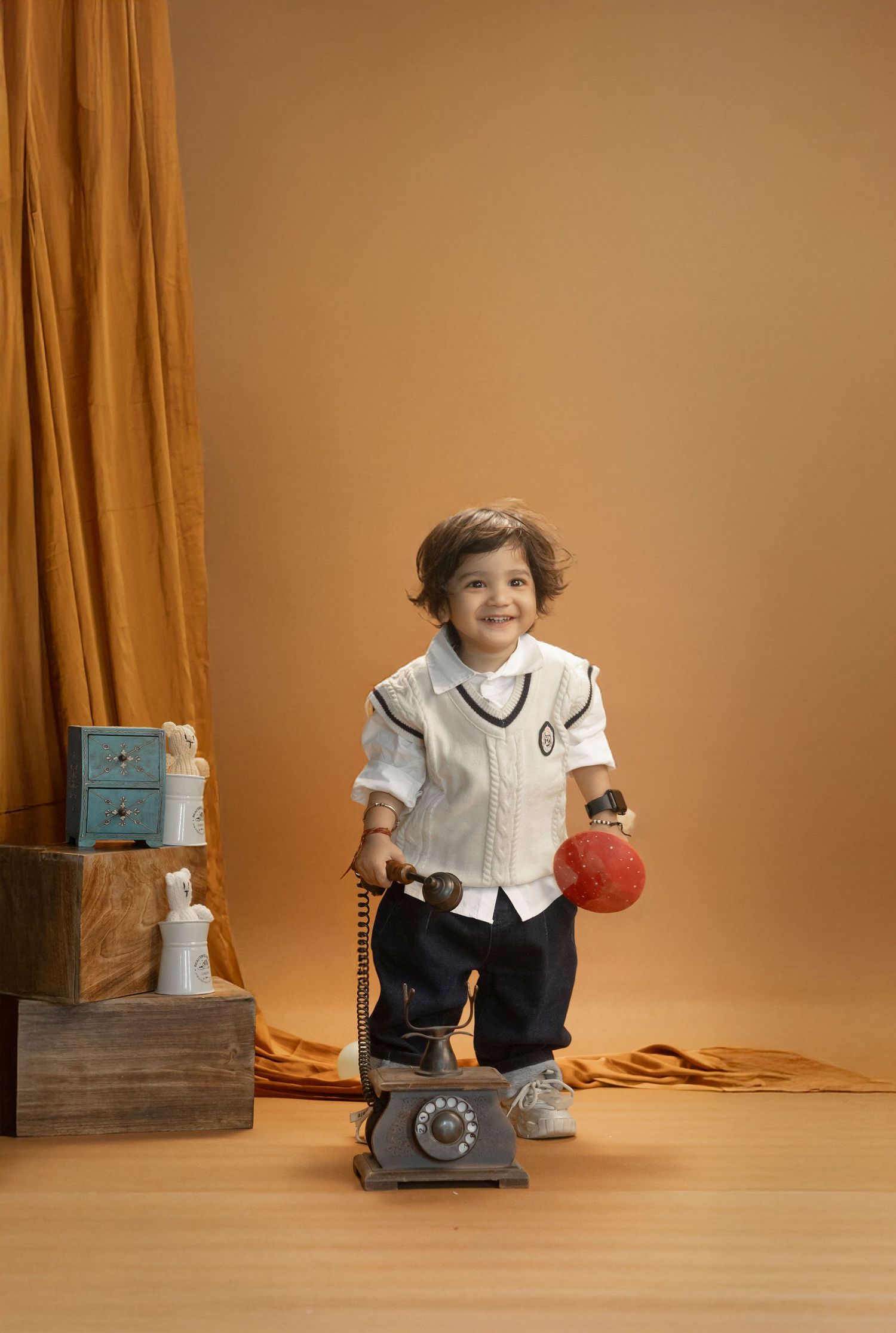 Smiling toddler in white shirt and navy pants posing with vintage telephone and red ball.