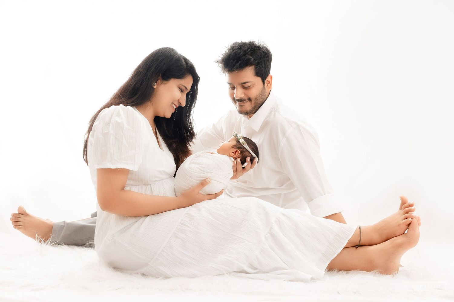Pregnant couple sitting on white floor, man smiling at woman holding baby shoes during maternity photoshoot.