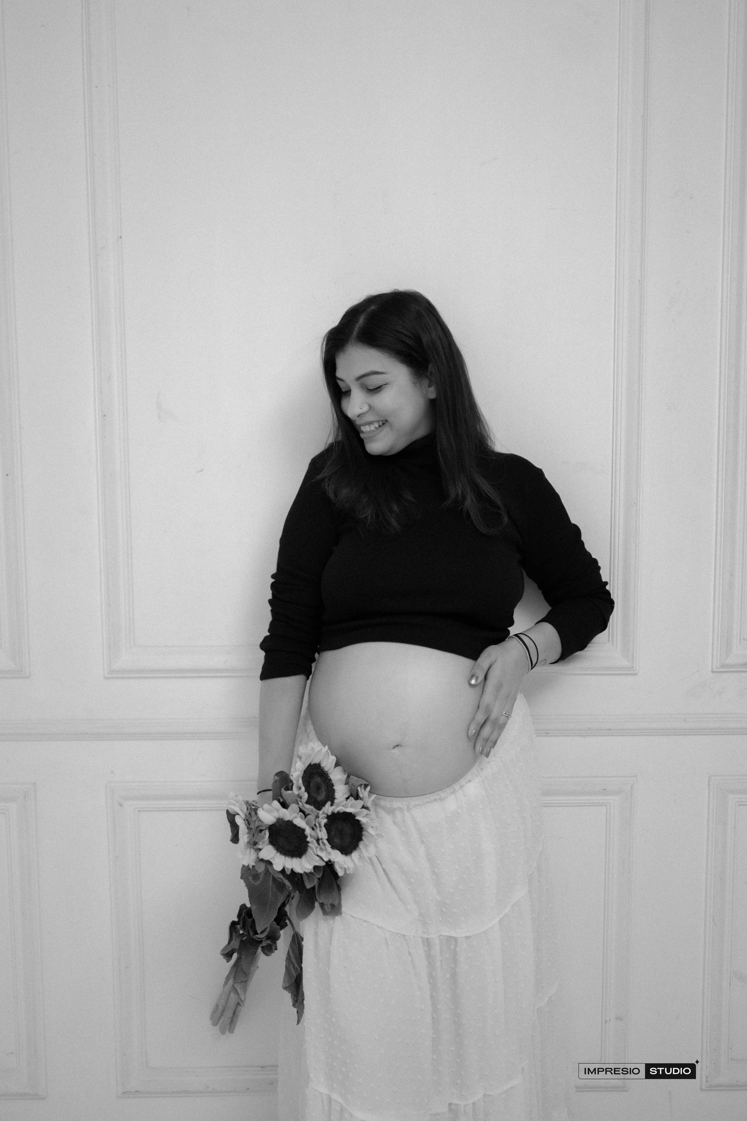 A smiling pregnant woman in black top and white skirt holds sunflowers in a black and white photo.