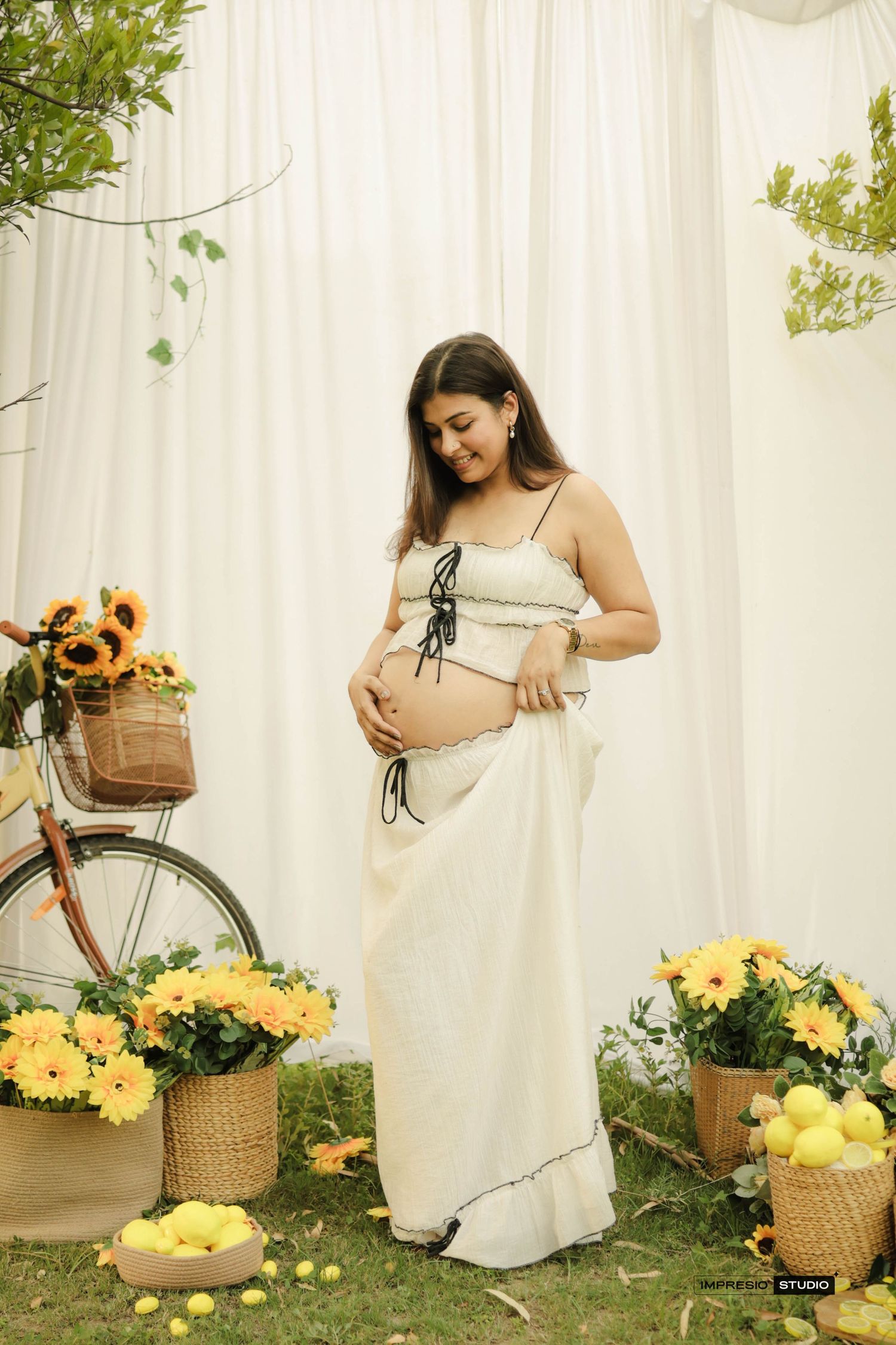 Pregnant woman in white crop top and skirt cradling baby bump, surrounded by yellow flowers and sunflowers outdoors.