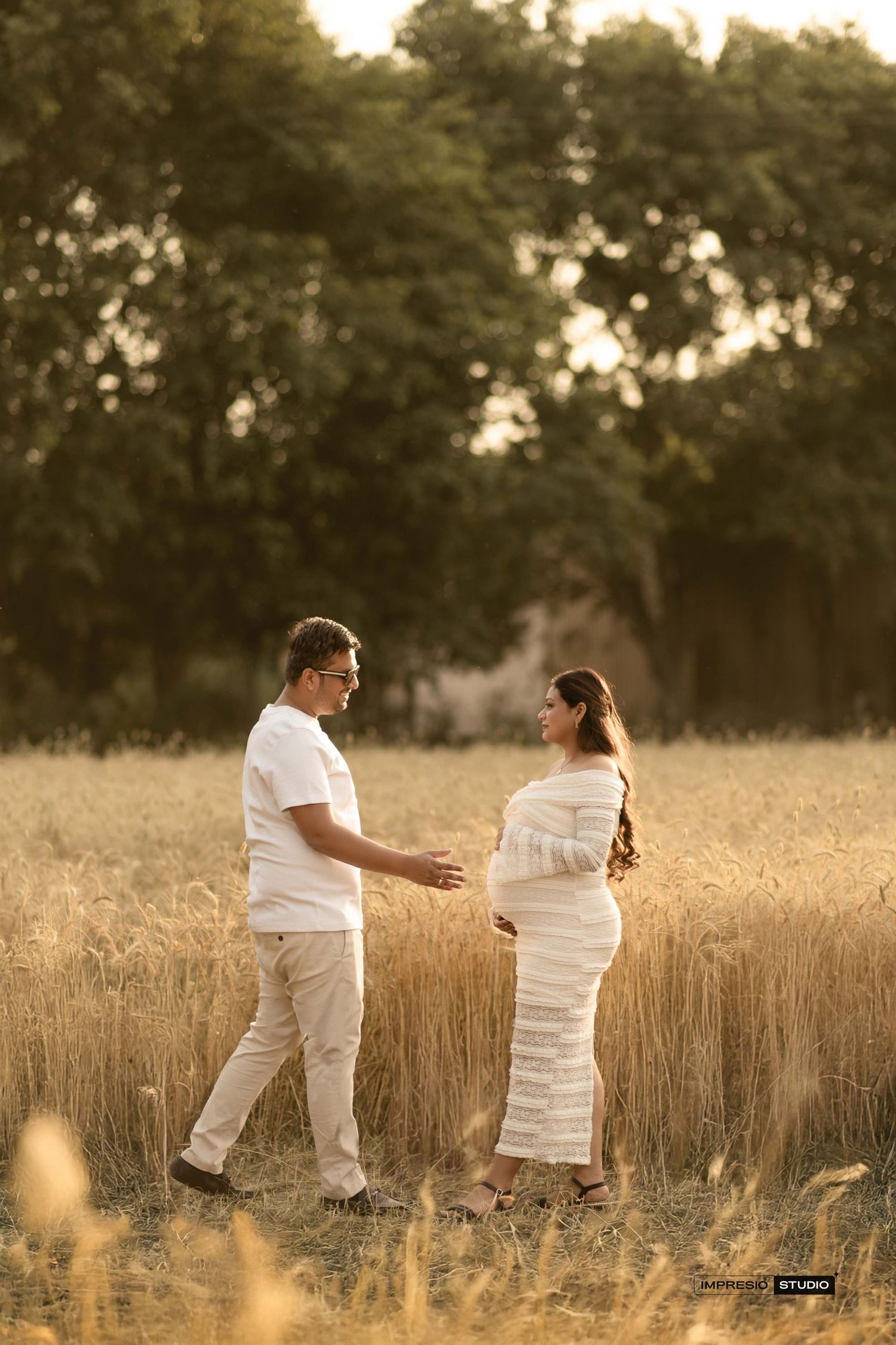 Couple in white outfits sharing a tender maternity moment in a golden wheat field at sunset.