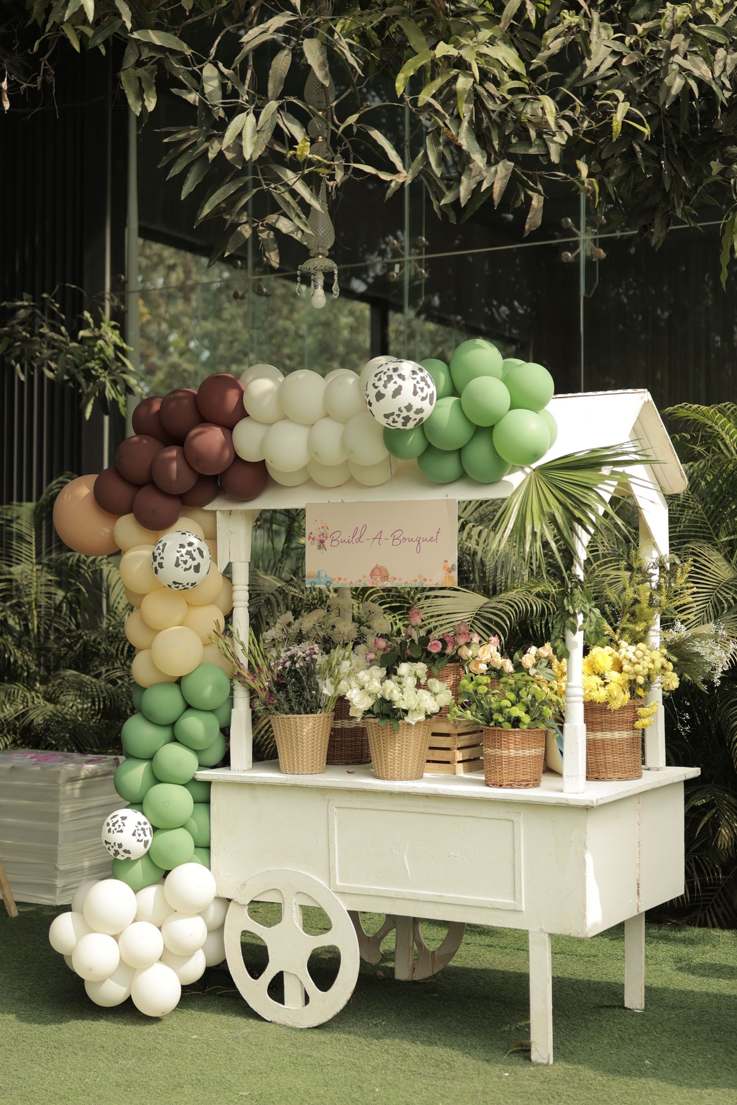White flower cart decorated with colorful balloon arch in green, burgundy, and white at an outdoor garden party.
