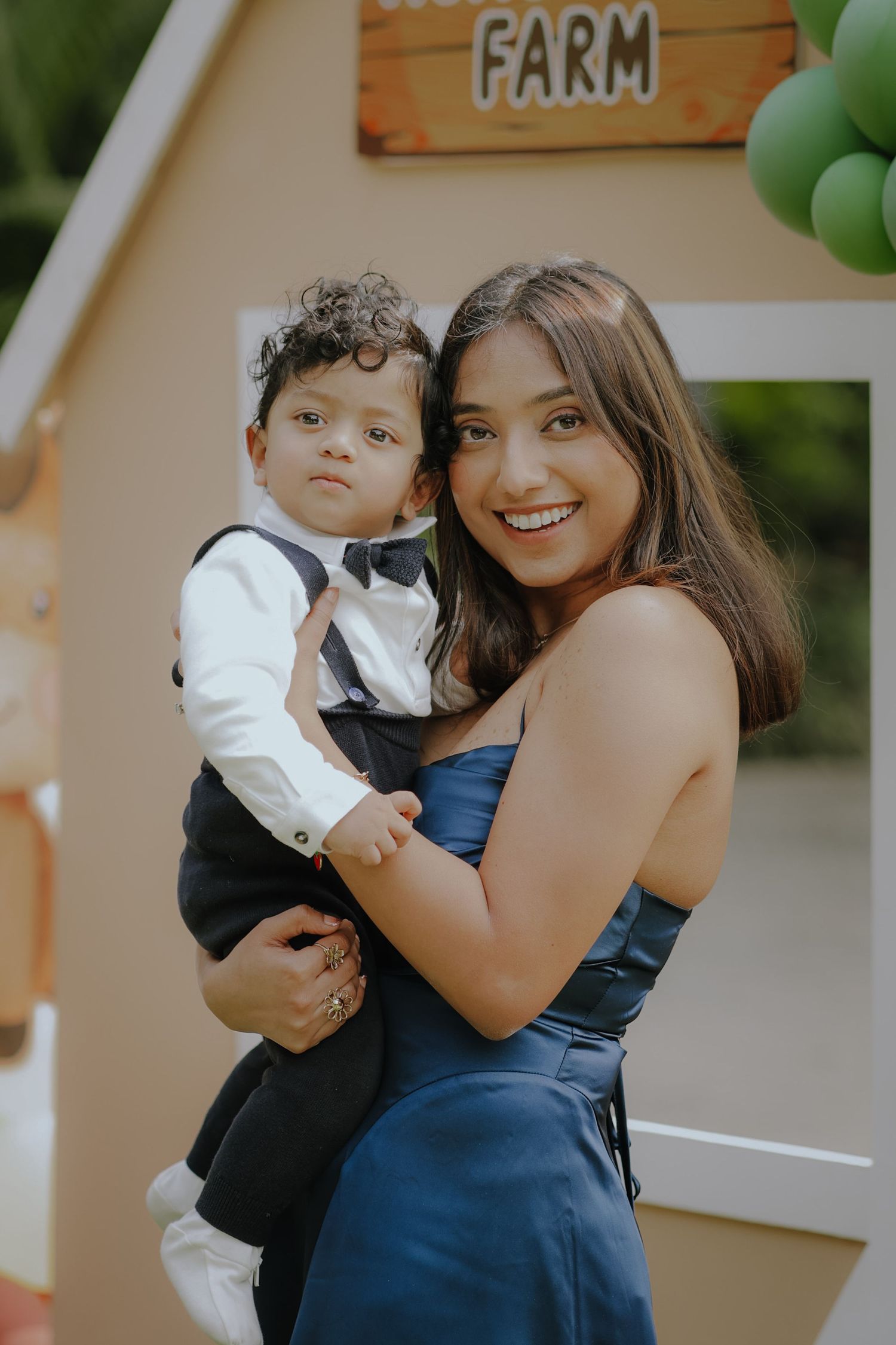 Young woman in blue dress smiling while holding a toddler boy in suit at a farm-themed birthday party.