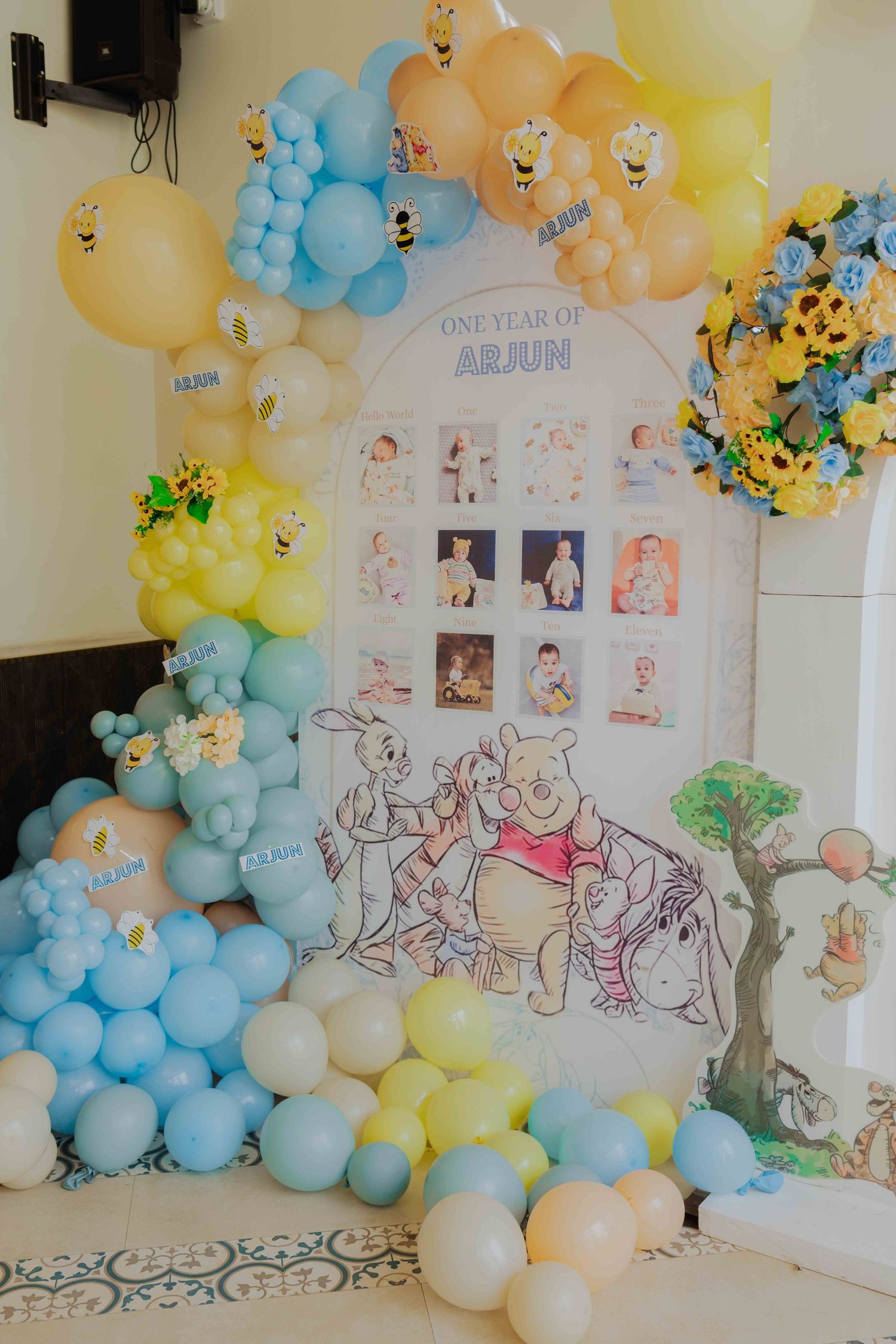 Winnie the Pooh first birthday party decoration with blue and yellow balloon arch and one-year photo display board.