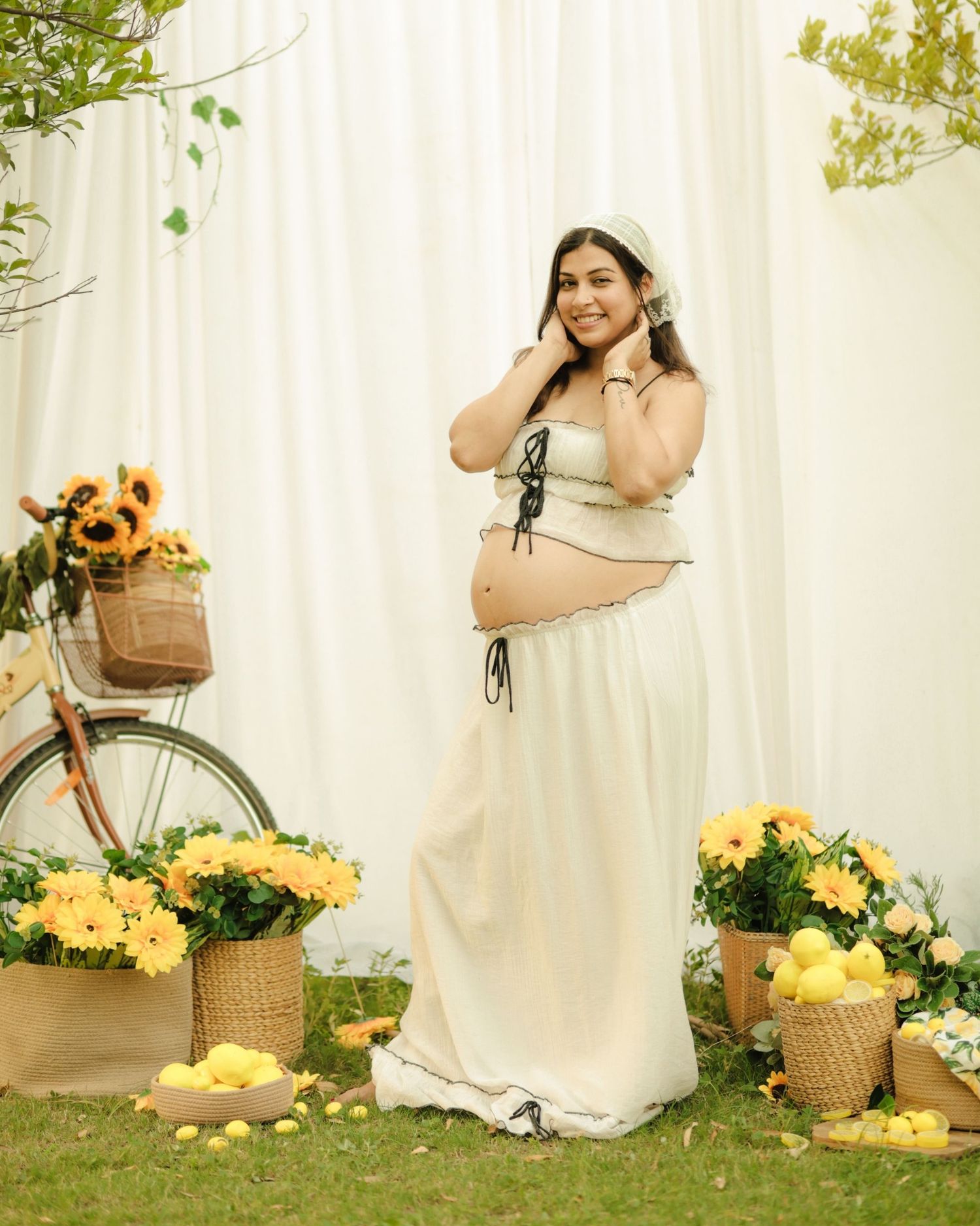 Pregnant woman in cream two-piece outfit poses among yellow flowers and sunflowers with vintage bicycle backdrop.