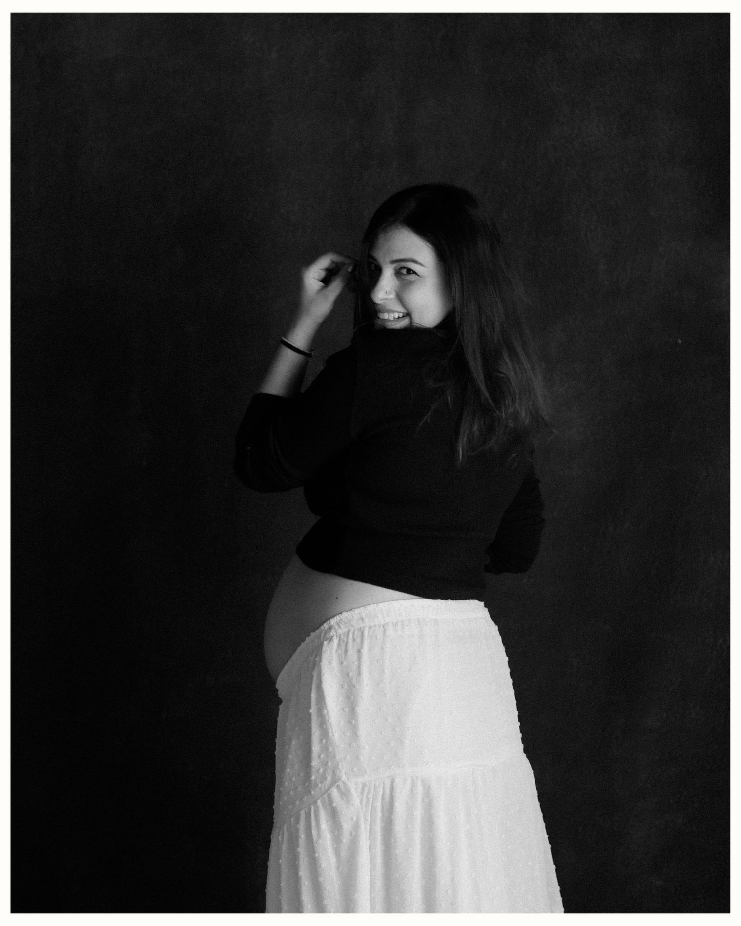 Black and white maternity photo of a woman in dark top and white skirt, smiling over shoulder against dark backdrop.