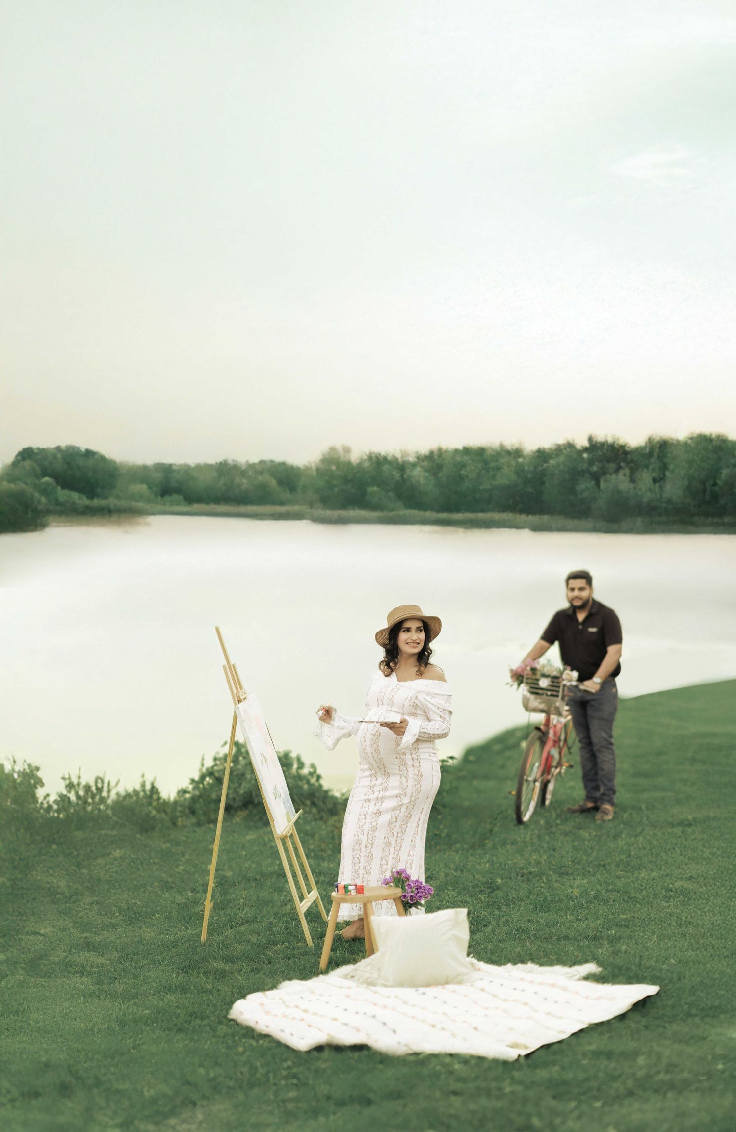 A pregnant woman in a white dress paints near a lake while a man with a bicycle stands behind her on green grass.
