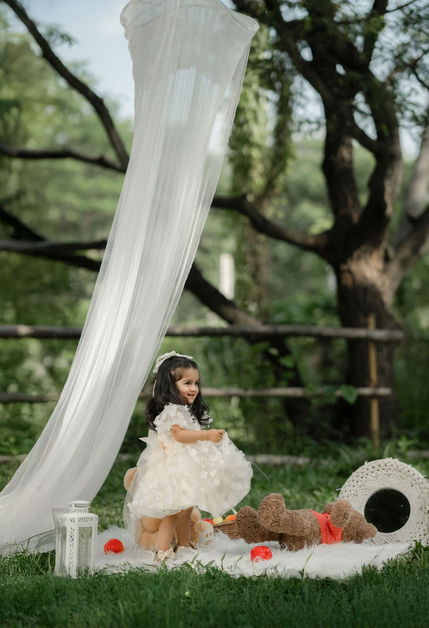 A little girl in a white dress plays with a teddy bear on a blanket outdoors near a flowing white canopy.