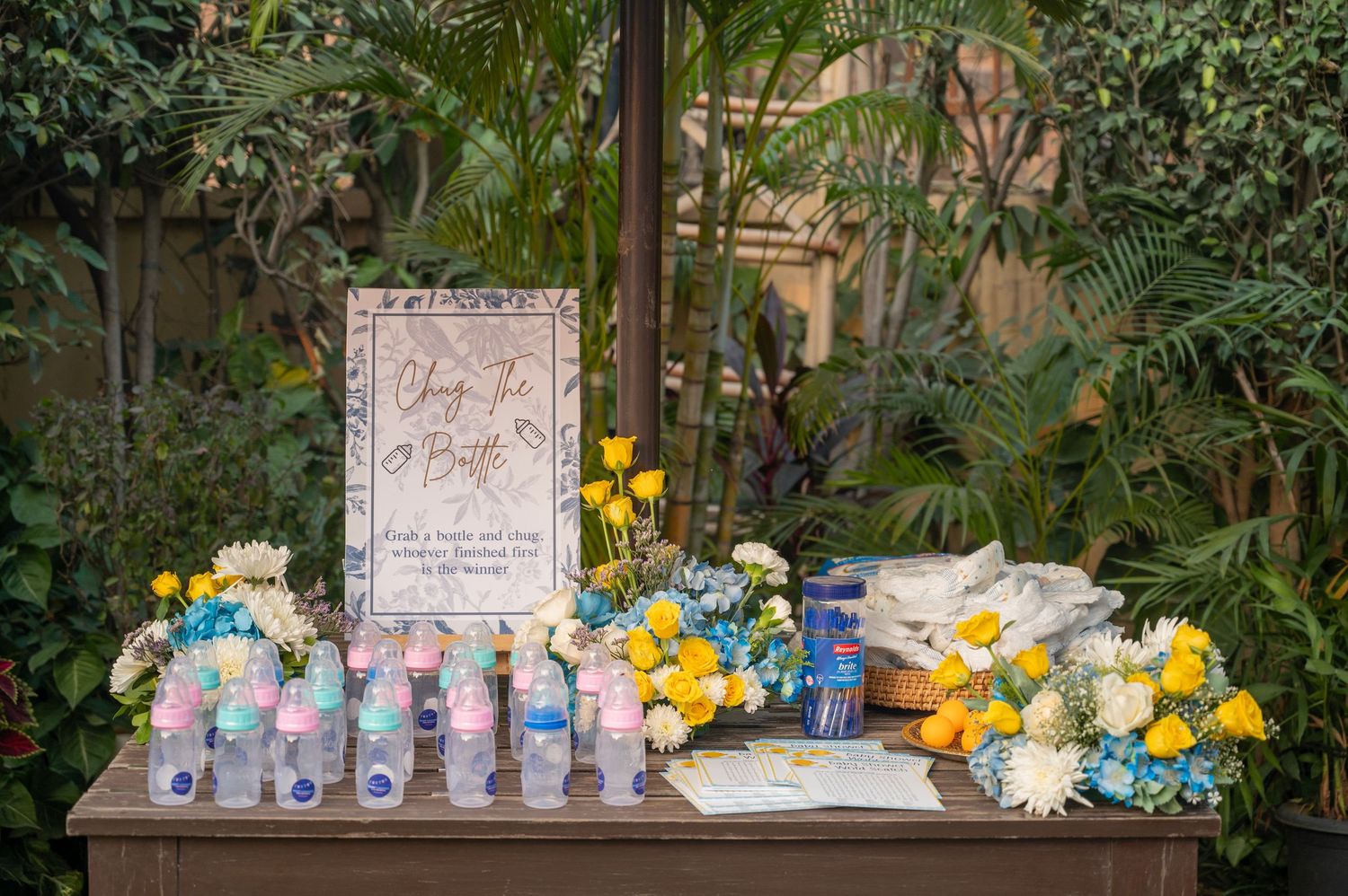 Wedding guest book table with floral arrangements, water bottles, and a decorative sign surrounded by lush greenery.