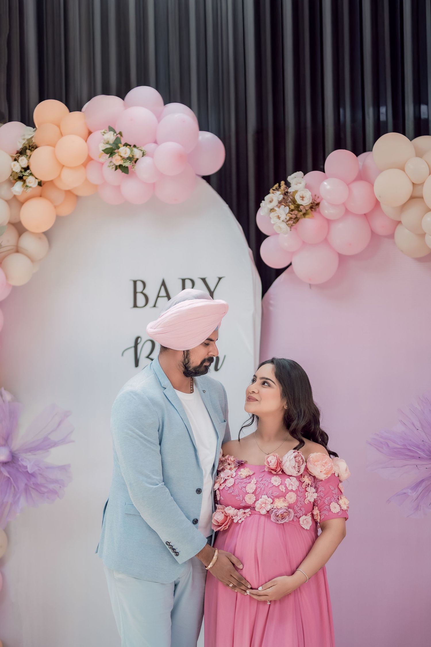 Couple at a pink baby shower with balloon arch, man in blue suit with pink turban, woman in floral pink maternity gown.