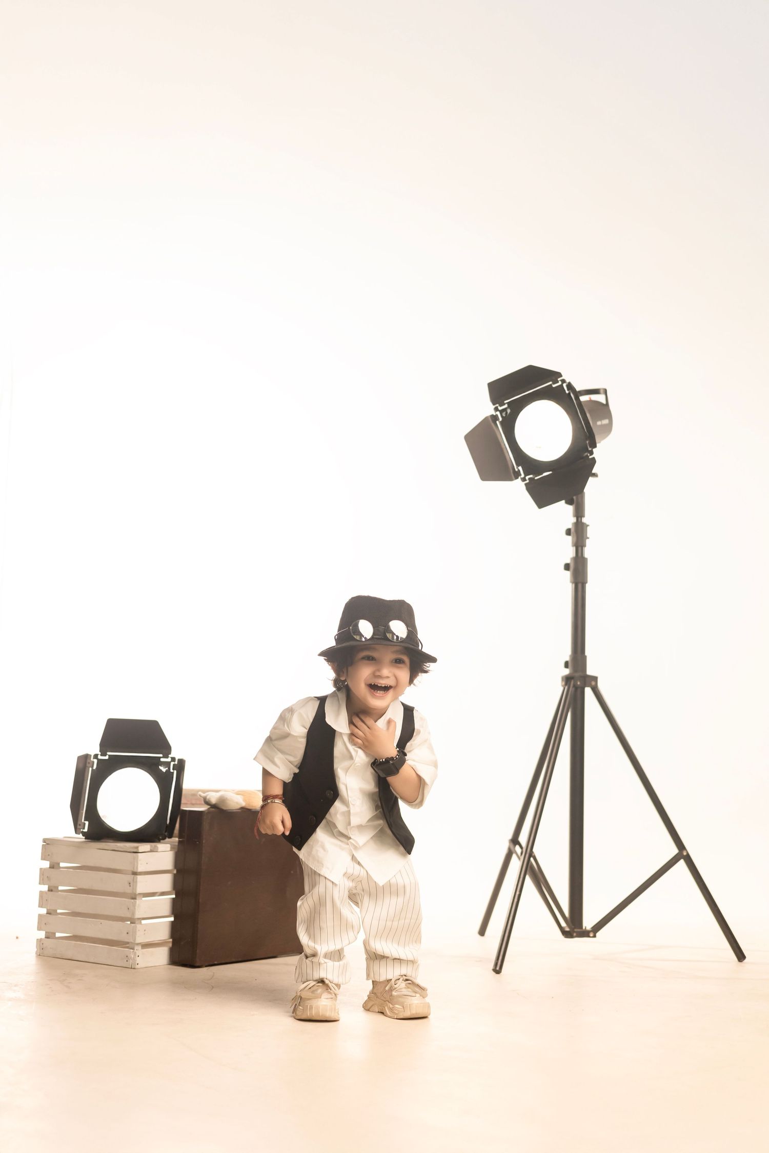A cheerful toddler in a striped suit and fedora poses with a vintage suitcase beside studio lights on white background.