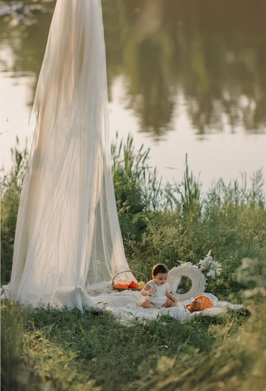 Baby sitting on a blanket by a serene lake, surrounded by orange pumpkins and a white flowing canopy.