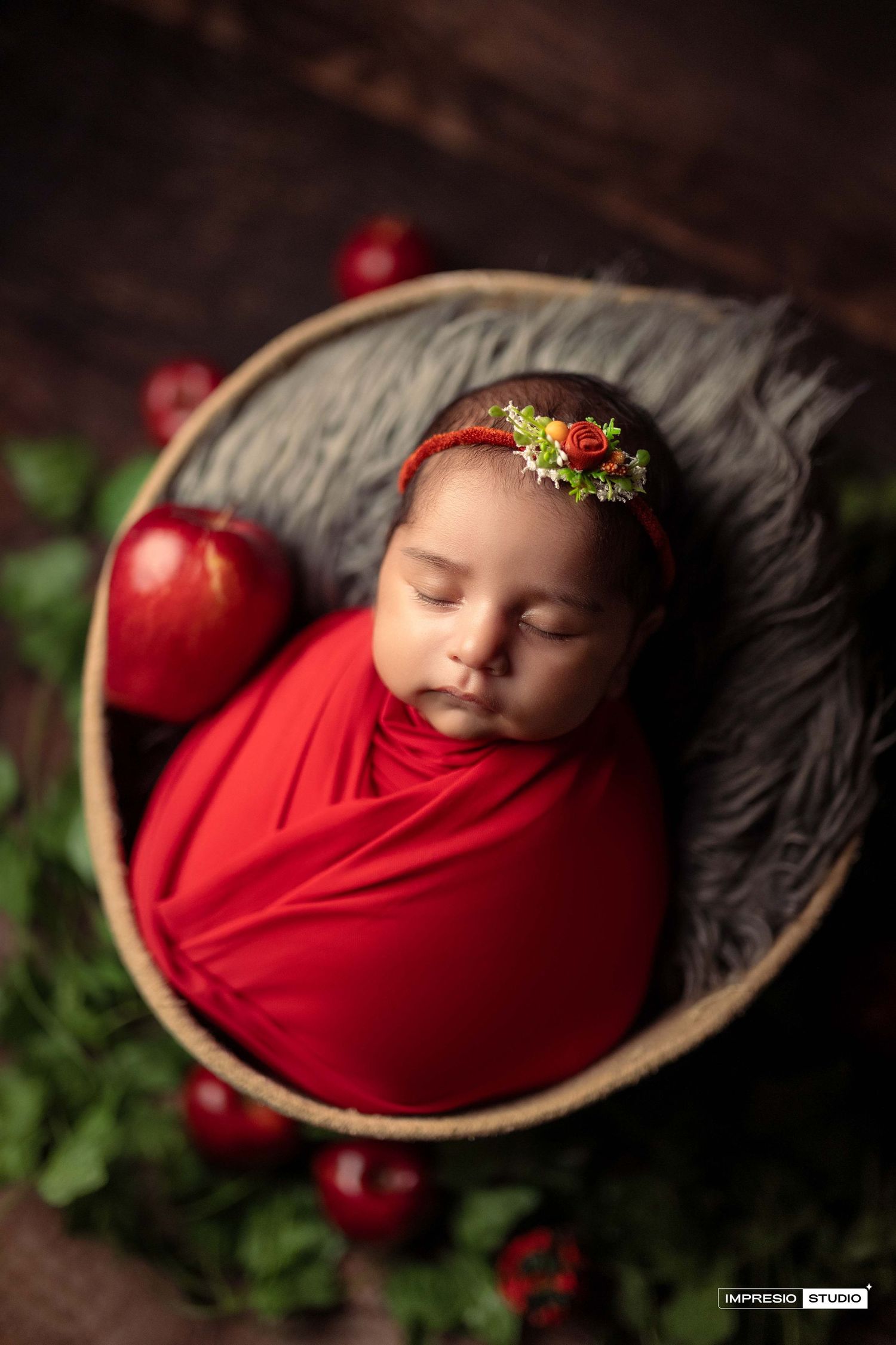 Newborn baby girl wrapped in red swaddle, sleeping in basket with floral headband and red apples, newborn photography.
