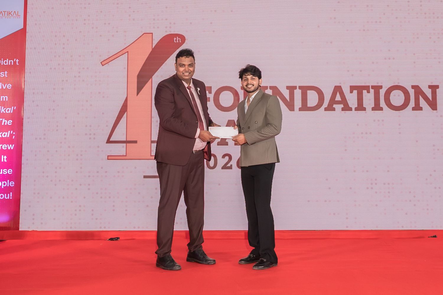Two men in formal attire shake hands while exchanging a cheque on a red-carpeted stage at a foundation award ceremony.