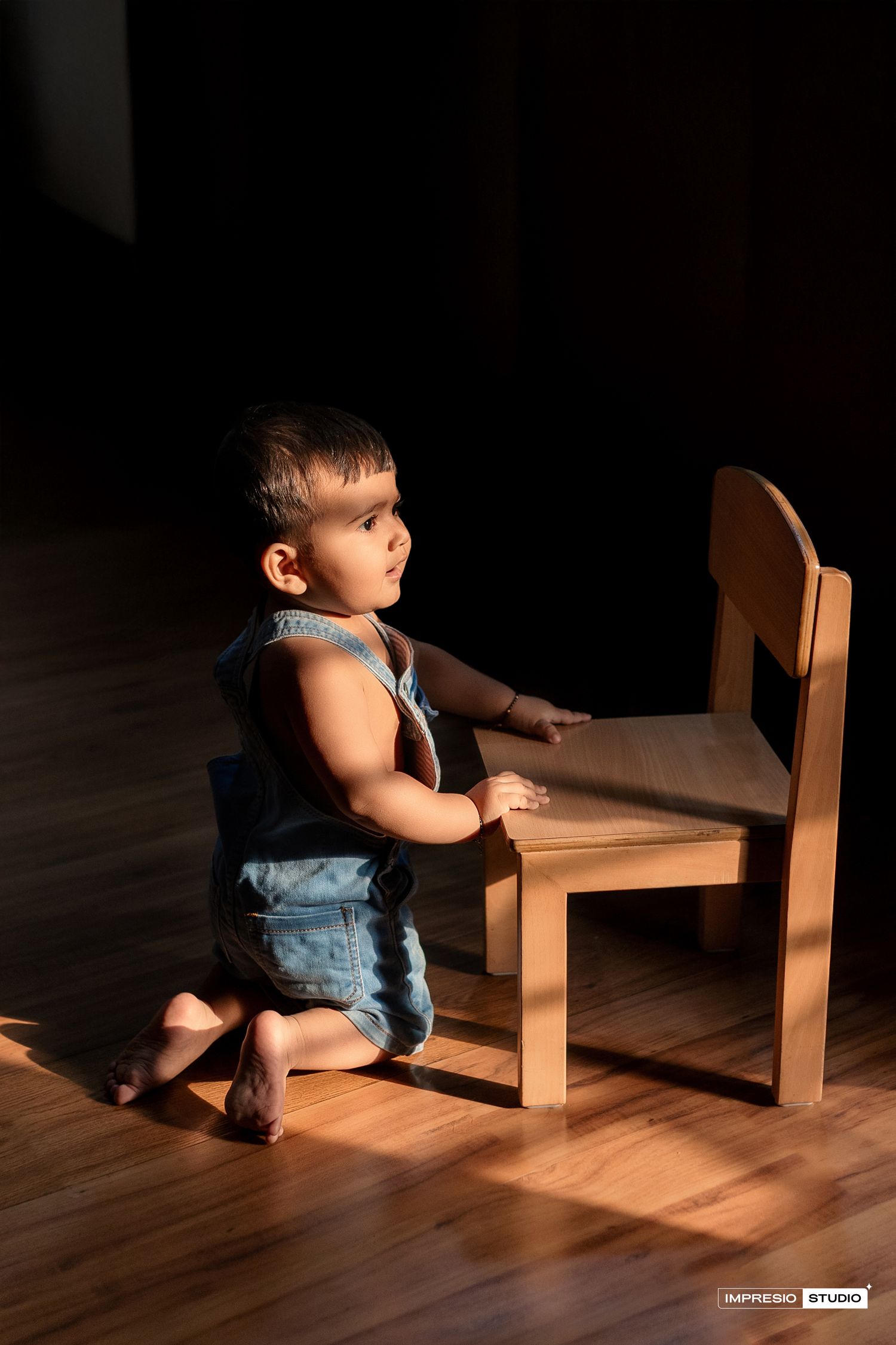 A small child in overalls sits on a wooden floor examining a wooden chair in dramatic lighting.