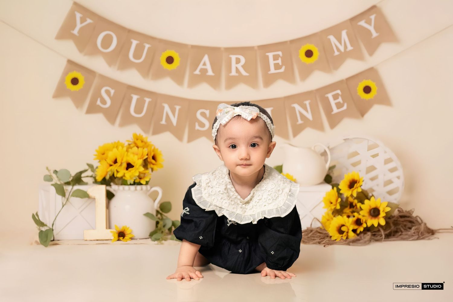 A baby in a black dress poses with yellow sunflowers and white roses against a beige backdrop with banner decor.