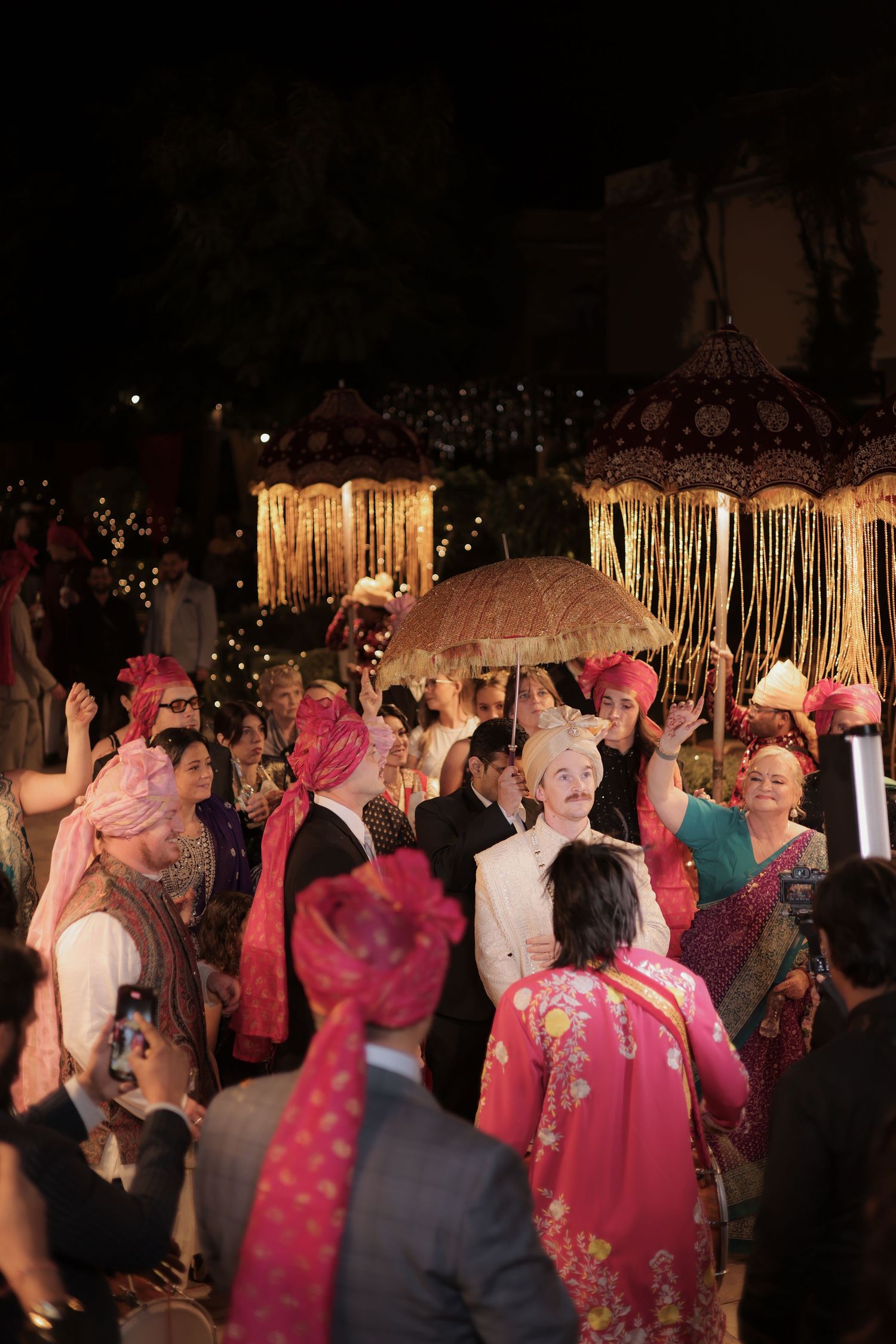 A lively nighttime celebration with people dancing in pink and red traditional outfits under decorative lighting.