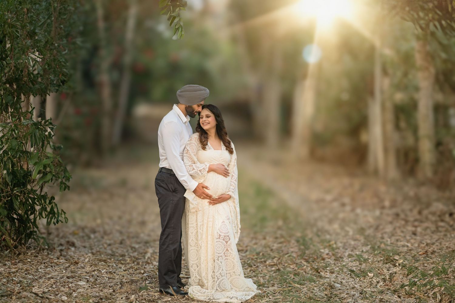 Romantic outdoor maternity photoshoot in white lace dress during golden hour in a wooded setting.