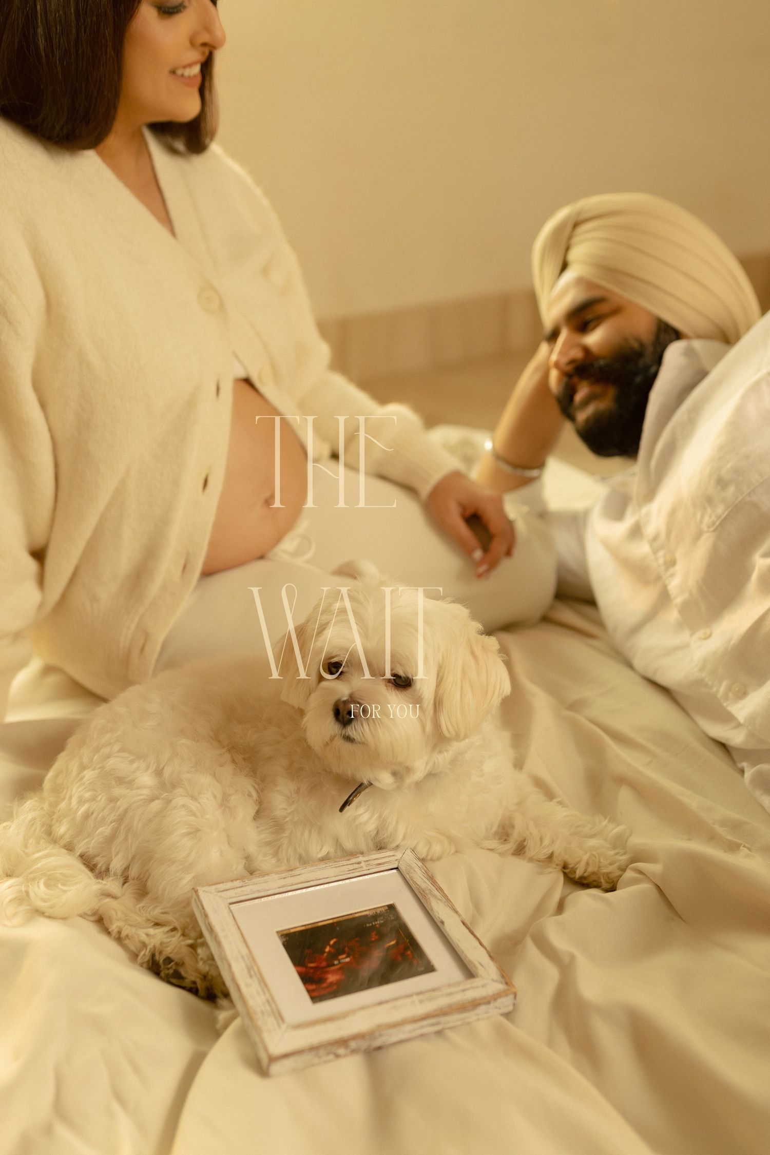 A couple relaxes on a white bed with their small white dog and a framed ultrasound photo.