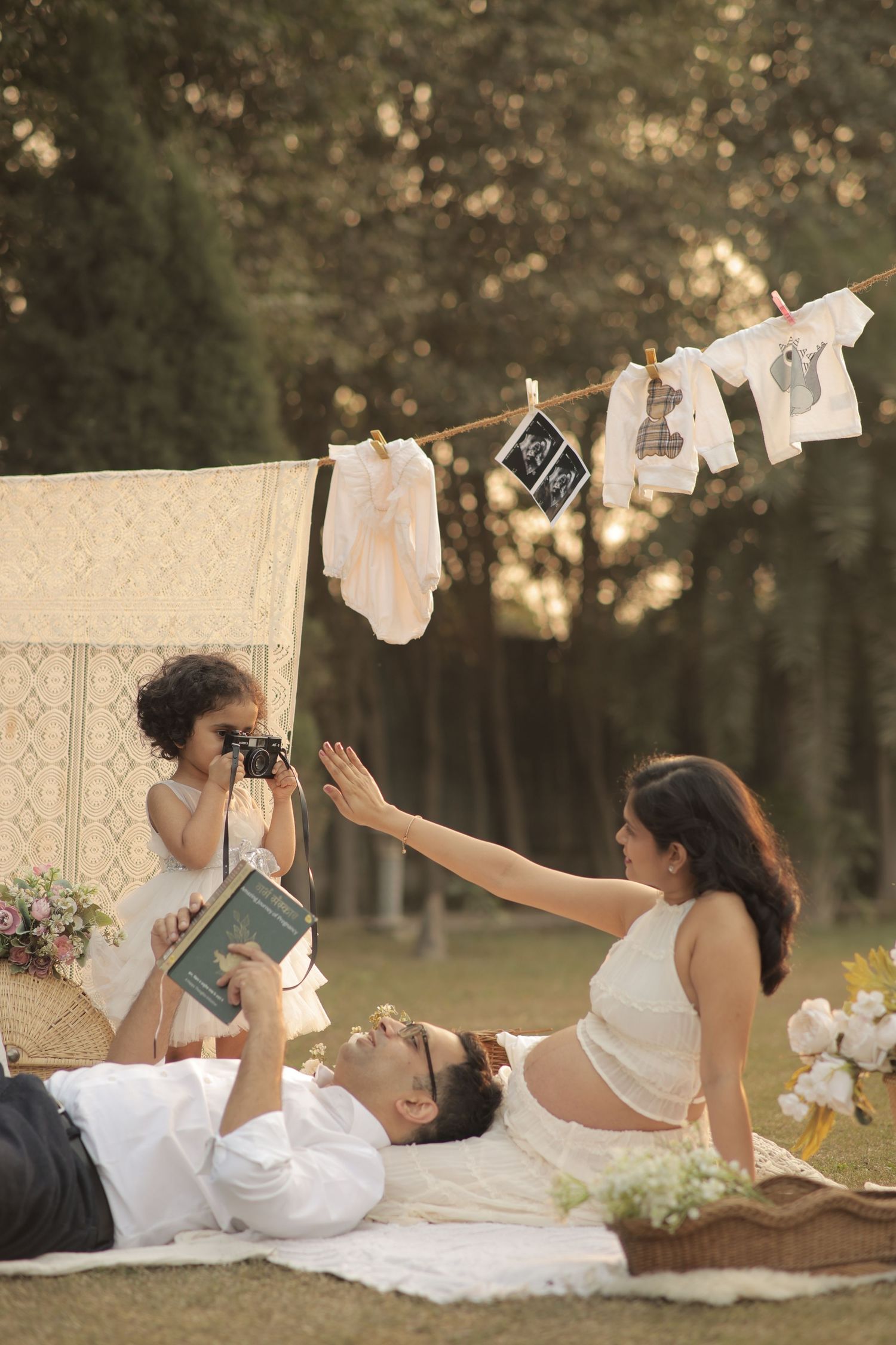 A family relaxes on a picnic blanket outdoors with baby clothes and ultrasound photos hanging on a clothesline above them.