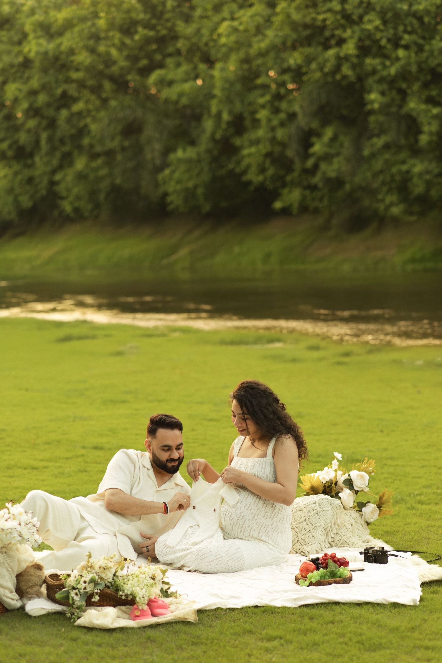 A couple in white outfits lies on a blanket having a romantic picnic in a lush green field with flowers.