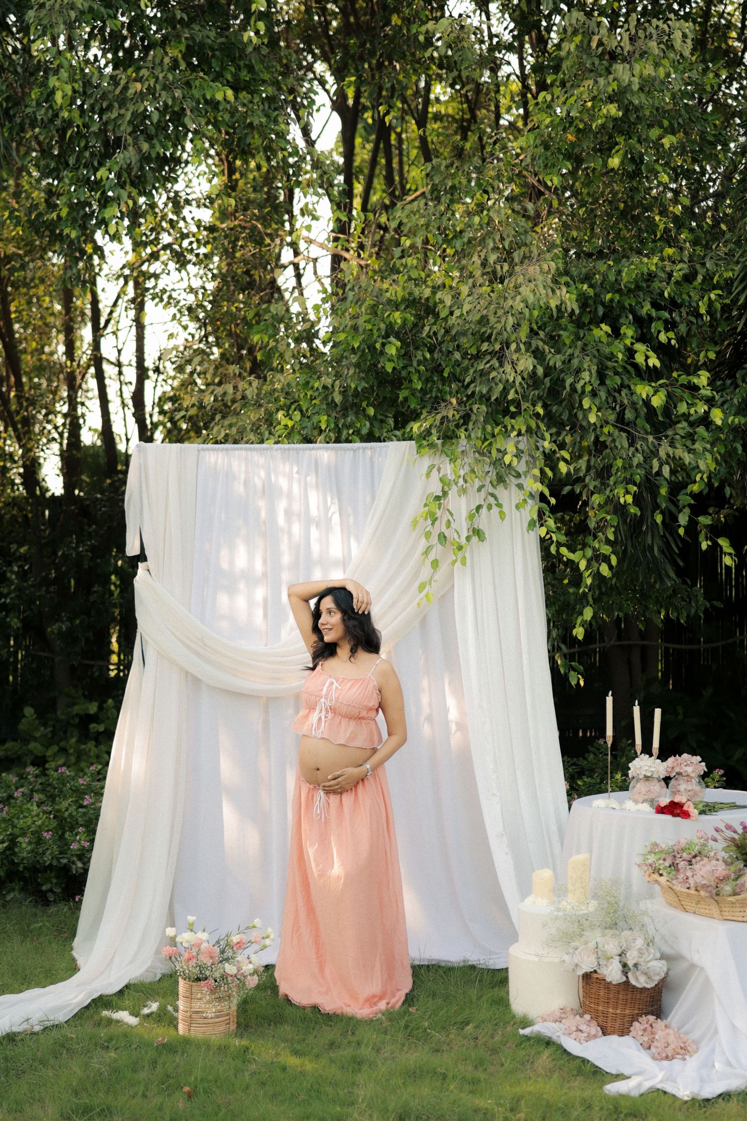 Pregnant woman in pink dress poses outdoors at elegant baby shower setup with white draping and floral decor.
