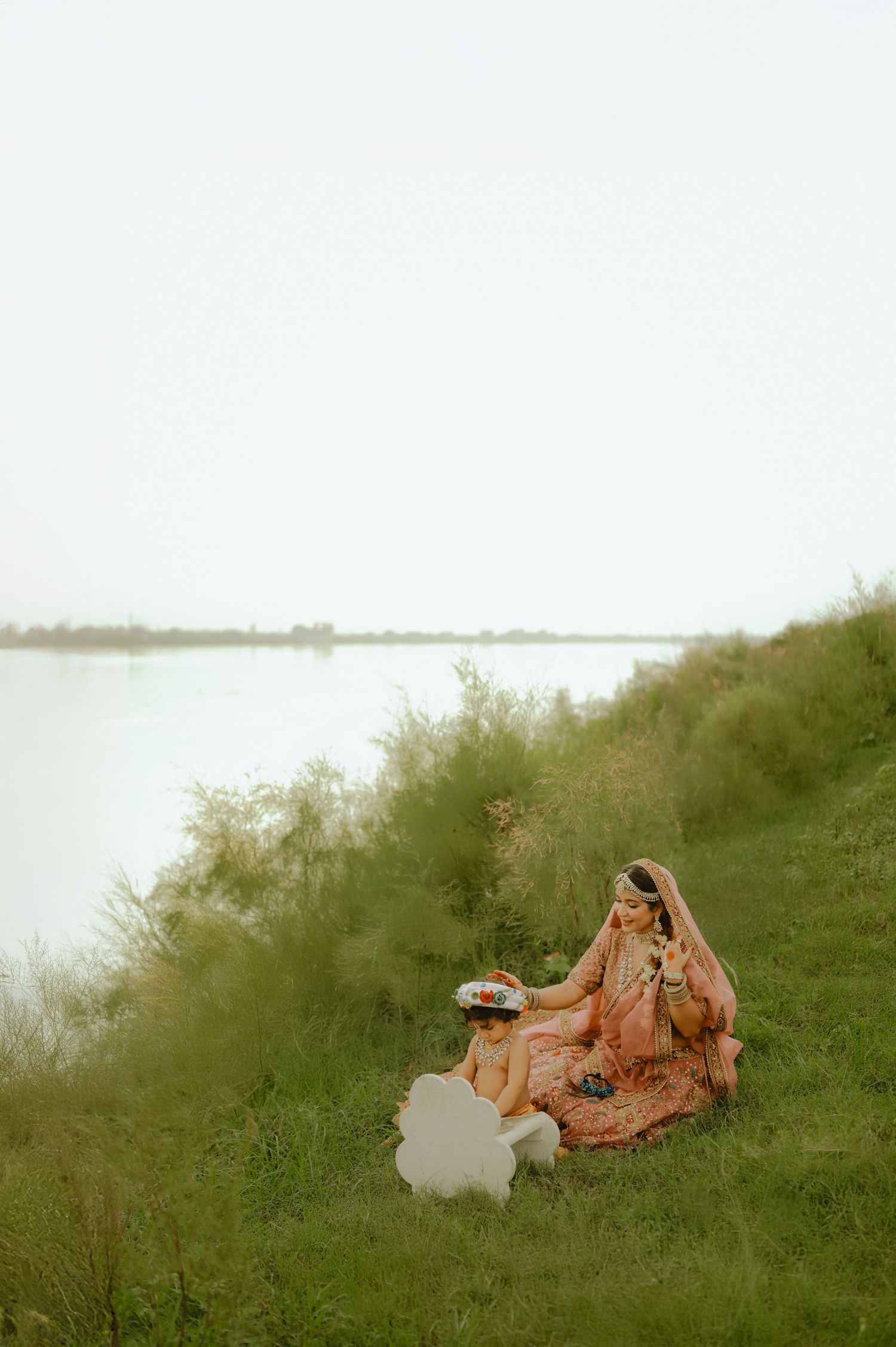 Bride in peach lehenga sitting on grassy riverbank, adorned with traditional jewelry, holding a decorative plate.