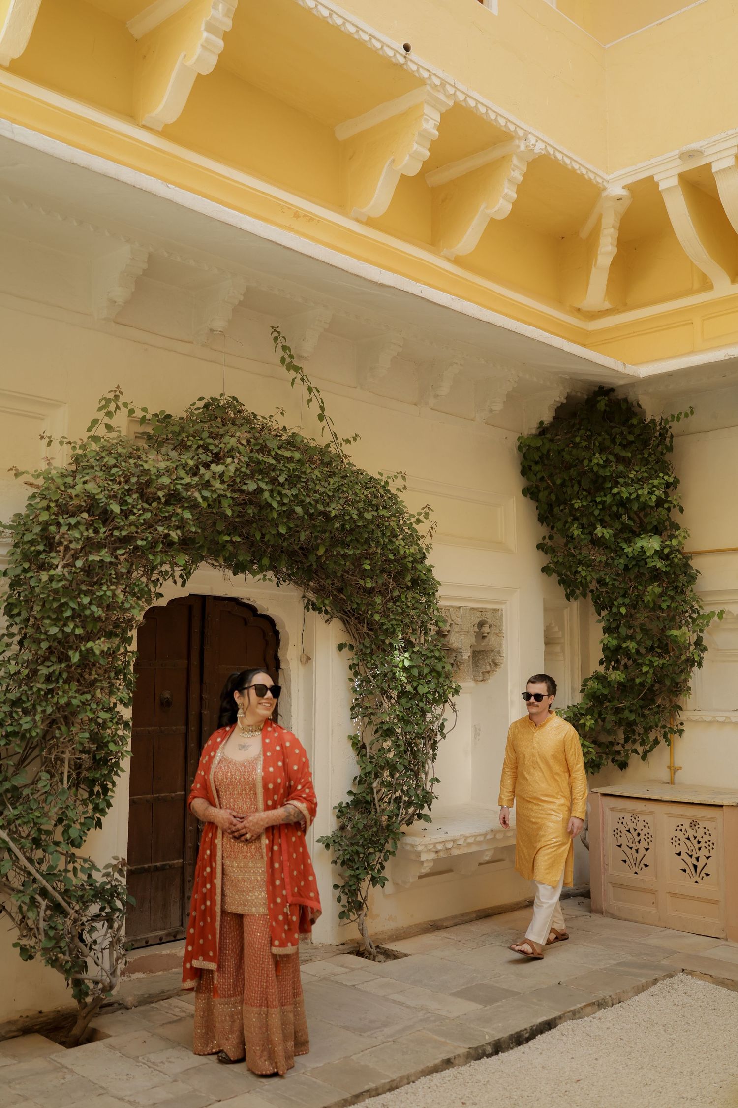 Two people in traditional Indian attire pose under ornate archways with climbing vines at a palace in warm yellow tones.