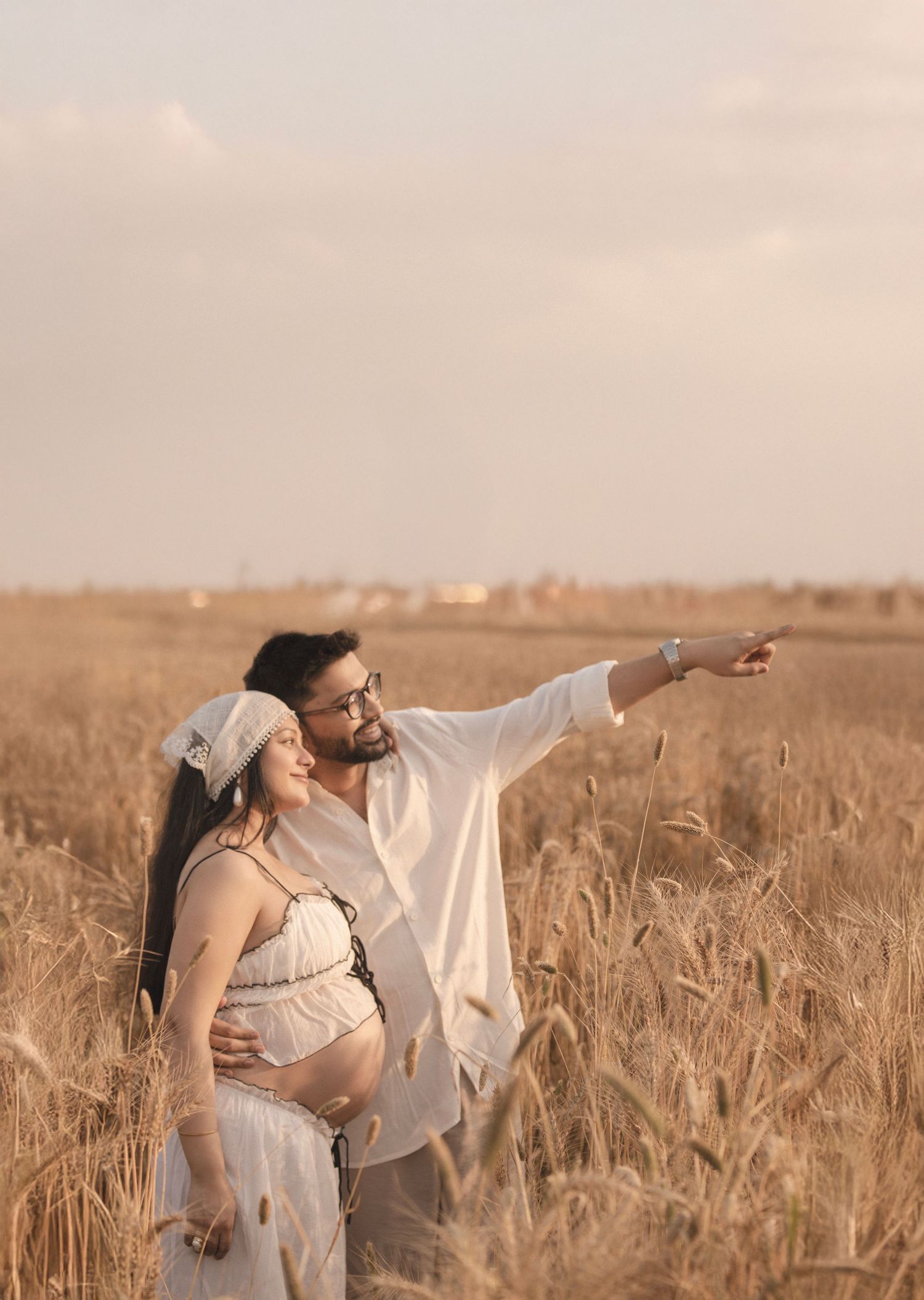 Couple in wheat field at sunset, man pointing into distance while embracing pregnant partner in white dress.