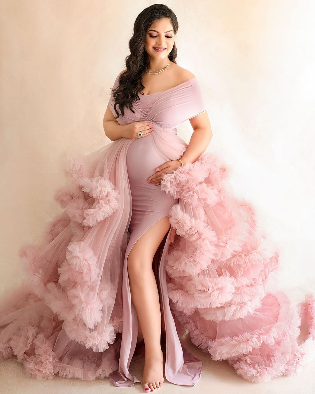 Maternity photo in pink tulle gown with dramatic ruffled train against beige backdrop.