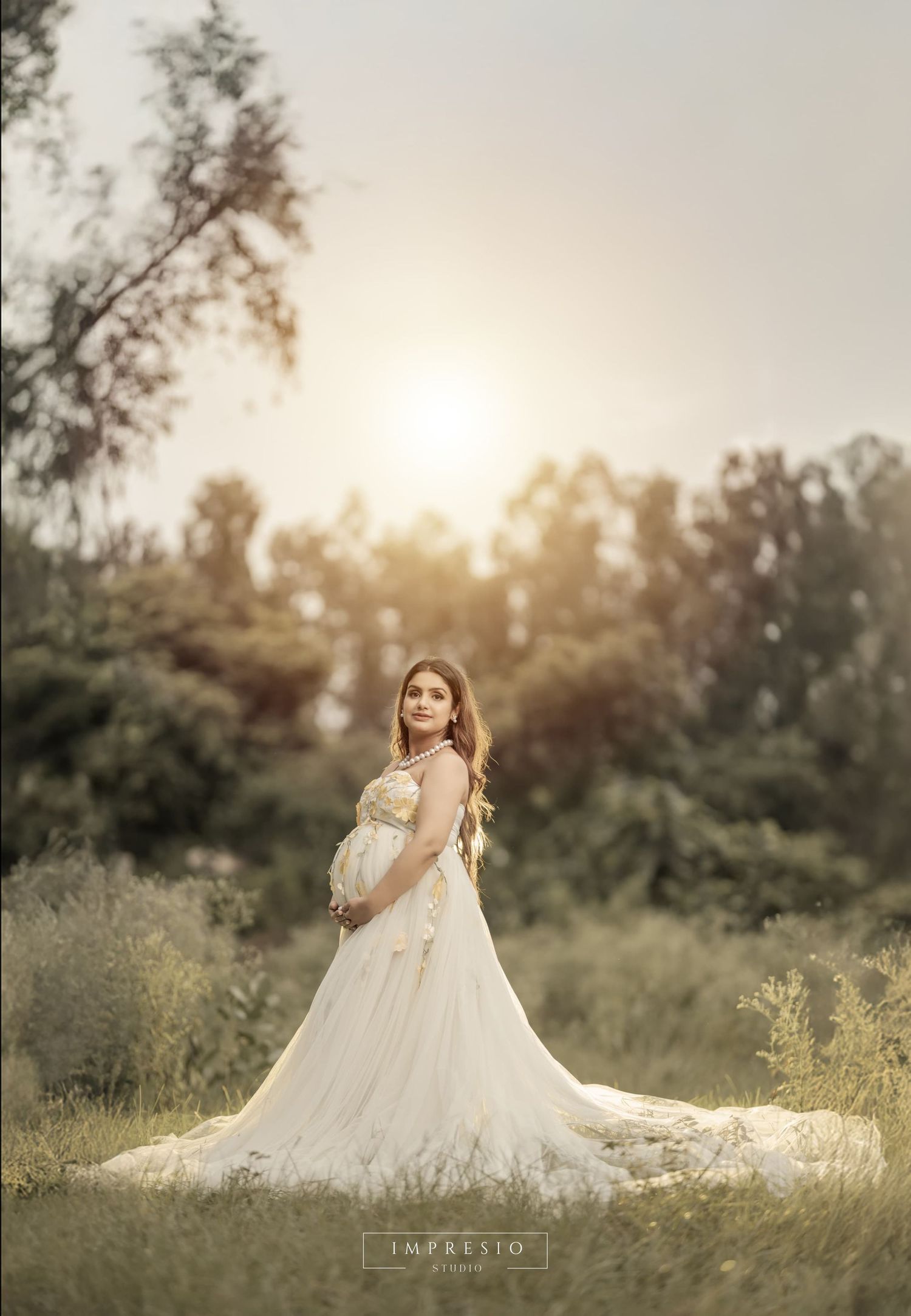 Elegant maternity portraits in a sunlit field with flowing white dress and natural outdoors setting.