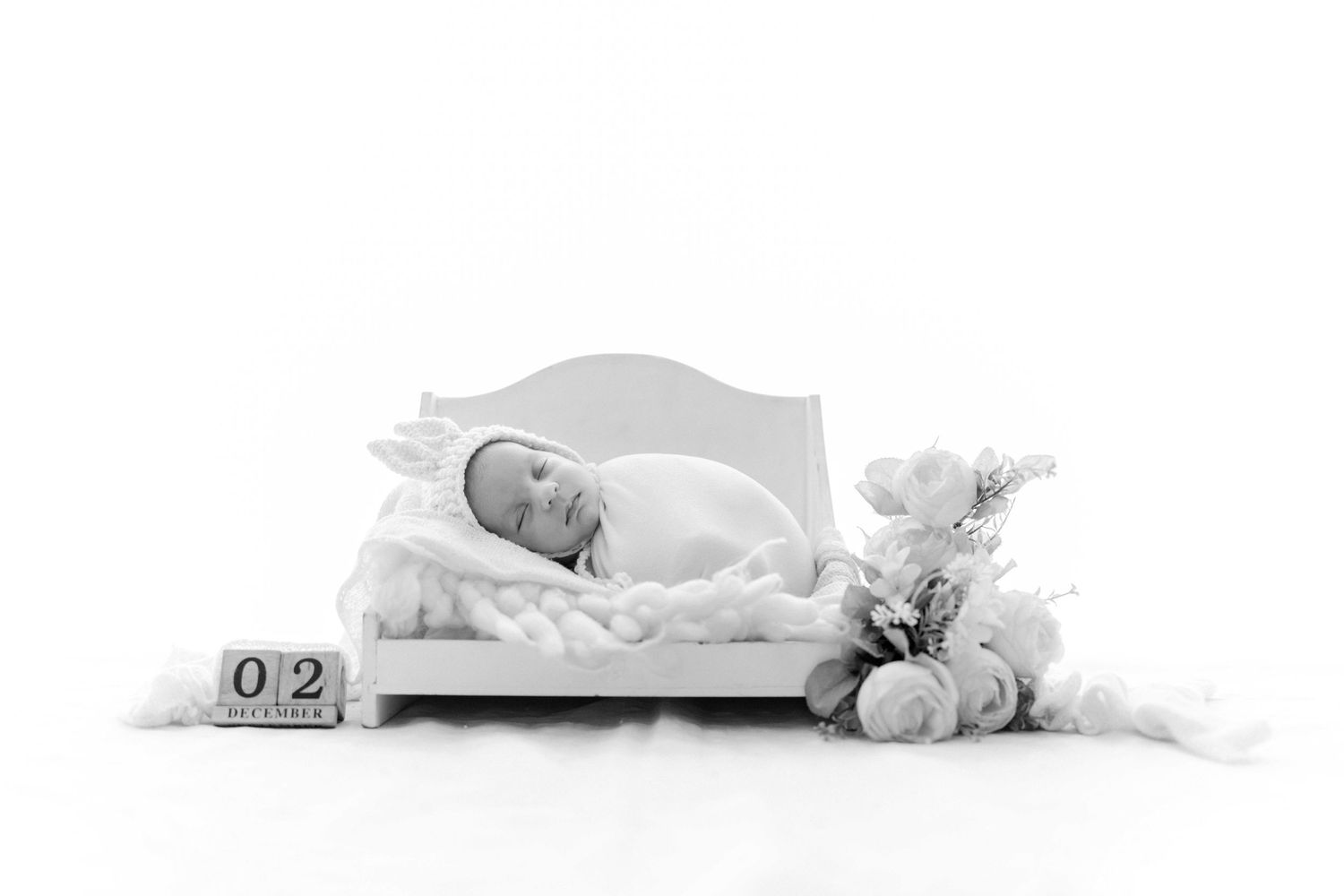 Black and white photo of sleeping newborn on white bed with date blocks showing 02 and floral decor.