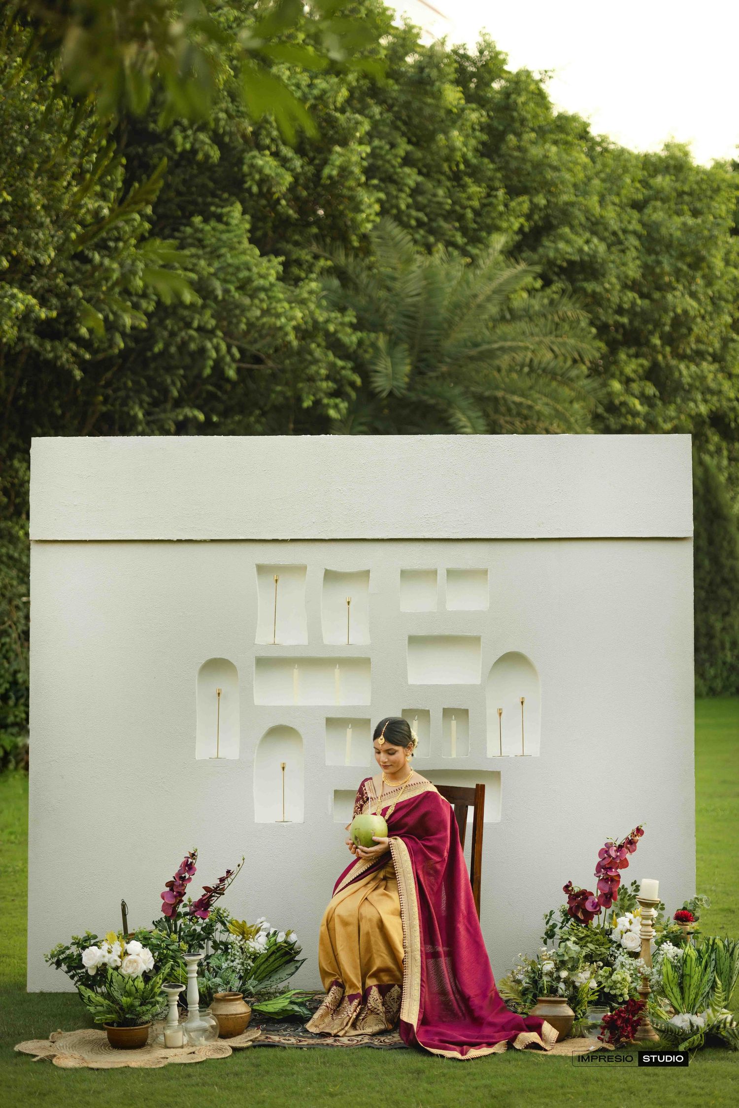 A woman in a maroon and gold saree sits gracefully before a white decorative backdrop surrounded by lush floral arrangements.