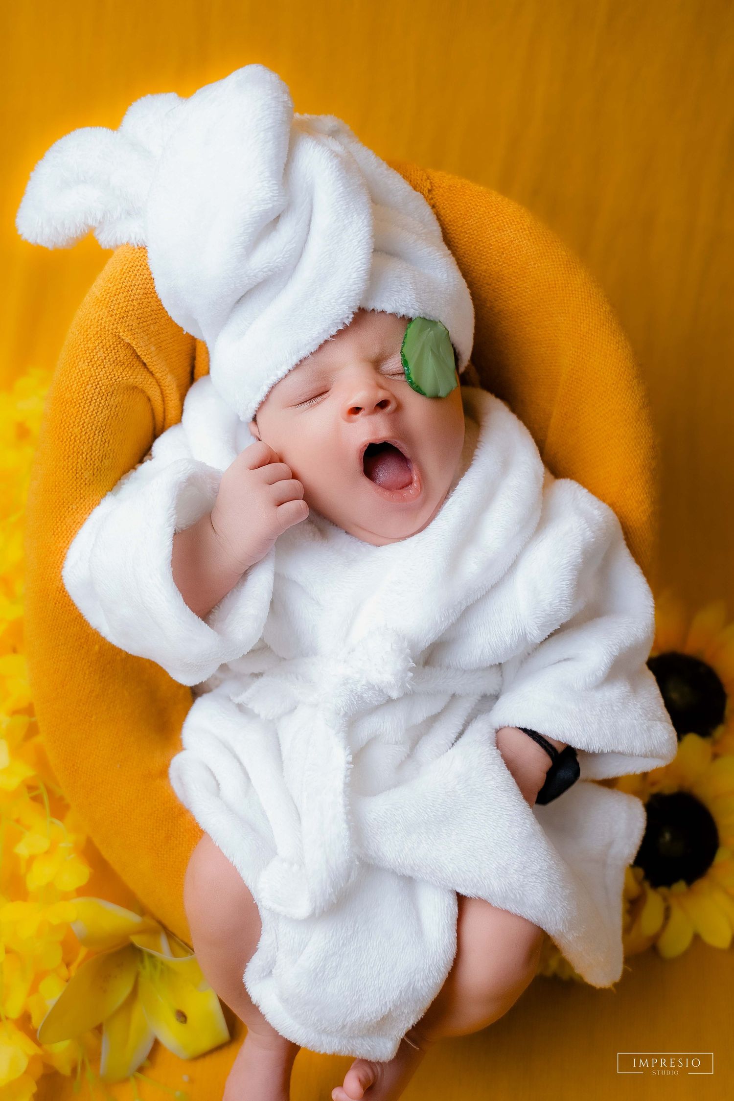 Newborn baby yawning in white fluffy robe and hat with cucumber eye patch on yellow background.