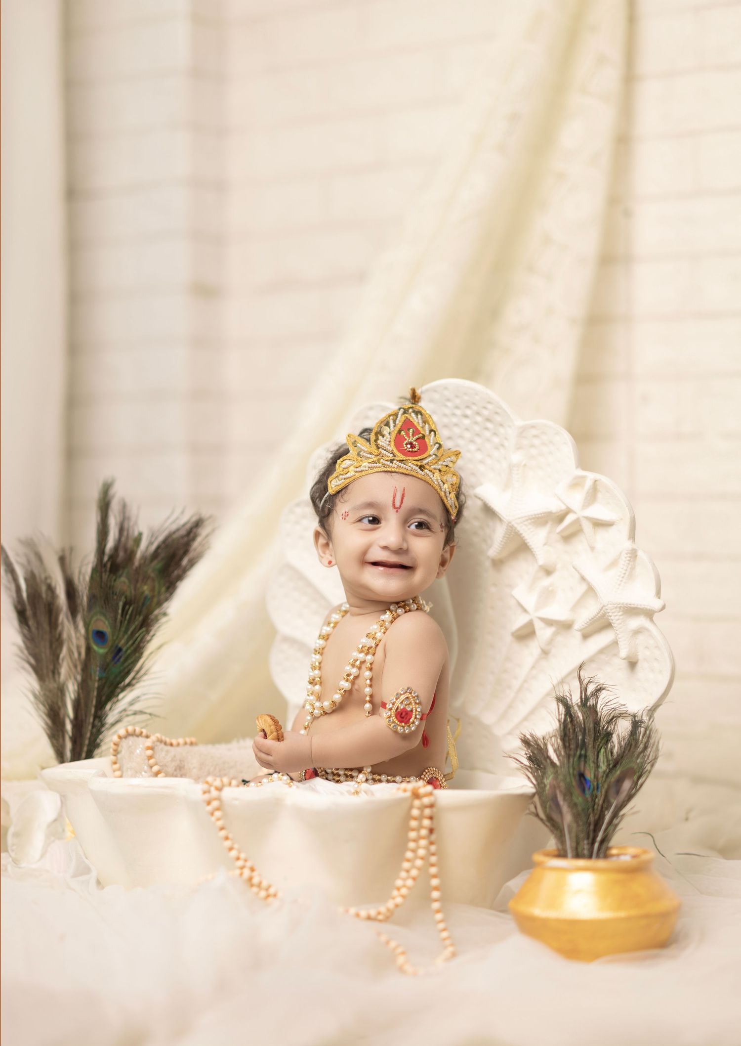 Baby dressed as Lord Krishna with crown, pearl necklaces, and peacock feathers in a white studio setup.