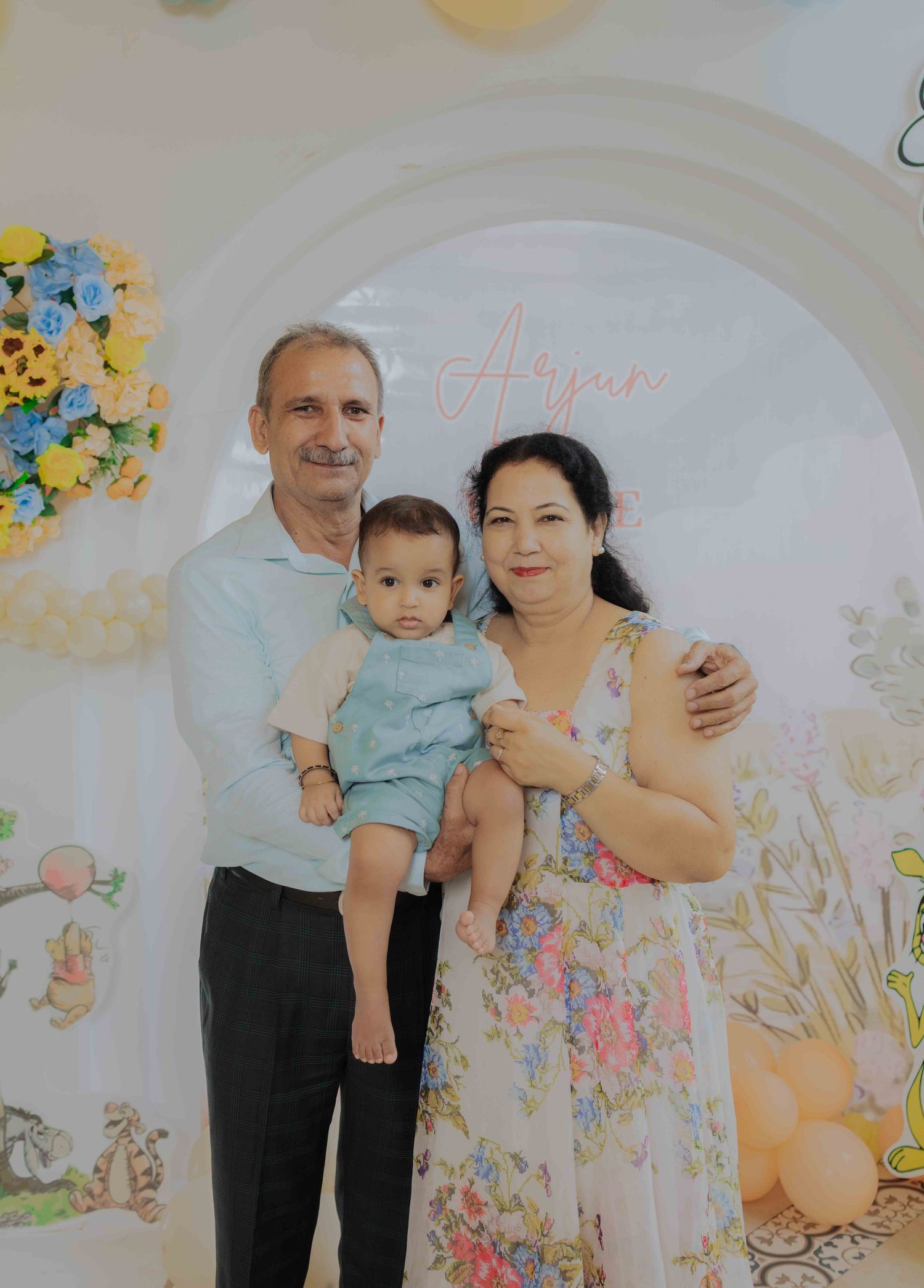 Family of three posing at a baby birthday party with floral arch backdrop and balloon decorations.