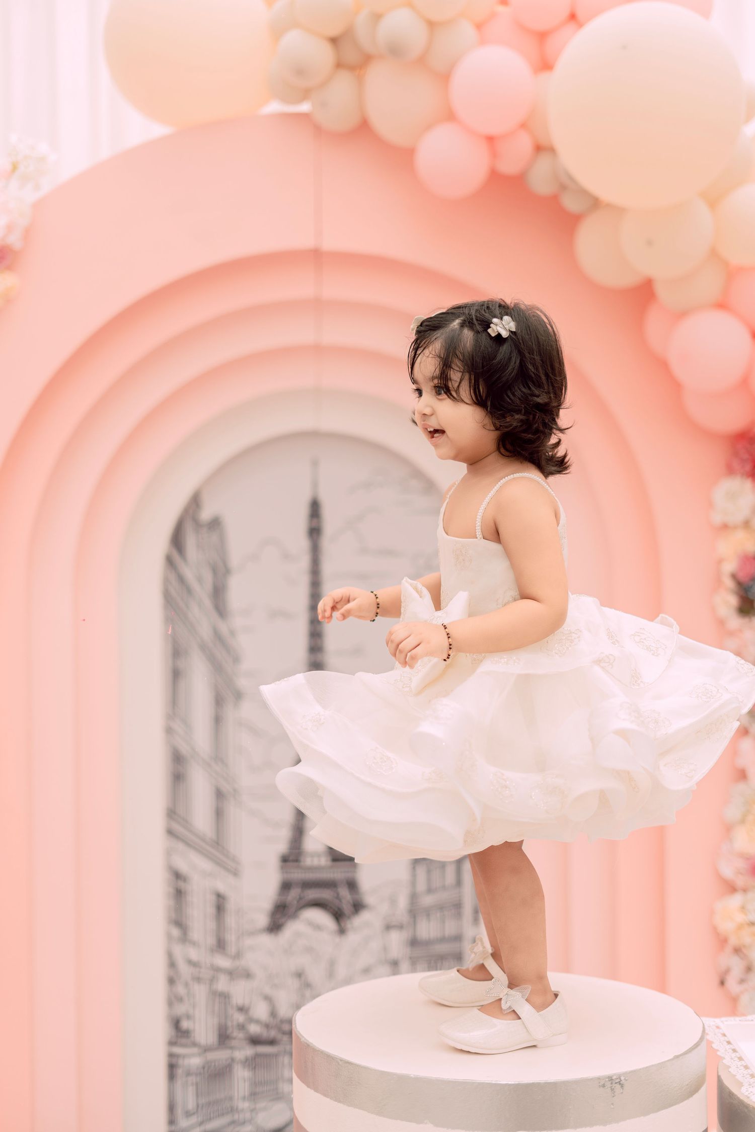 Toddler girl in white tutu dress standing on pedestal at Paris-themed birthday party with pink balloon arch.