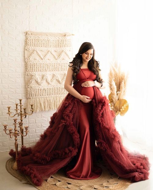 Flowing burgundy maternity dress with dramatic tulle overlay in a romantic studio setting with macrame backdrop.