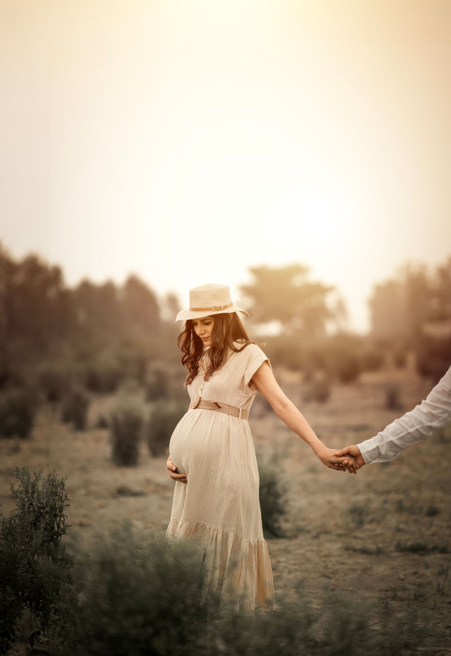 A pregnant woman in a flowing beige dress and sun hat holds hands with someone at sunset in a rustic field.