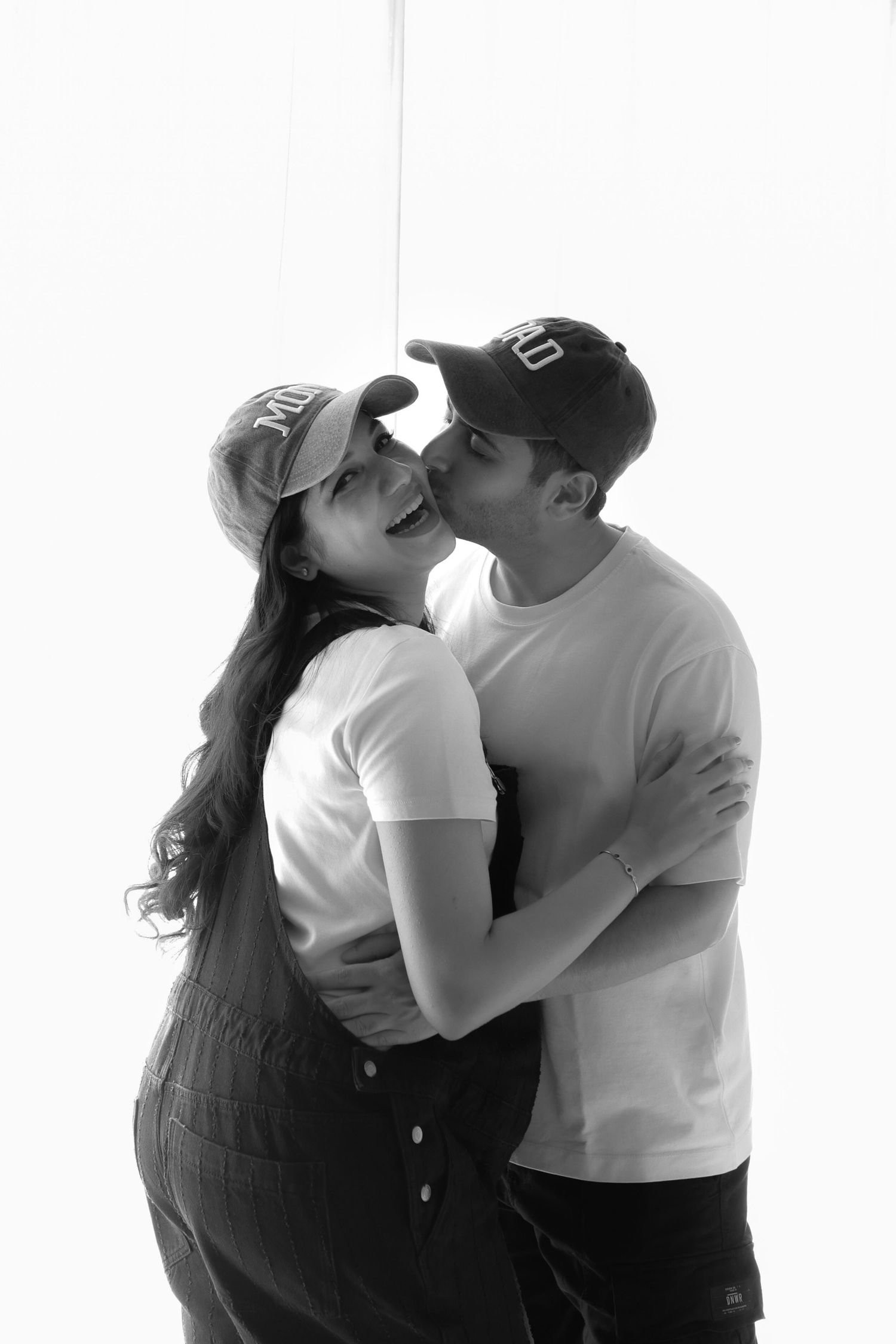 Black and white photo of a couple sharing a kiss, both wearing baseball caps and white t-shirts.