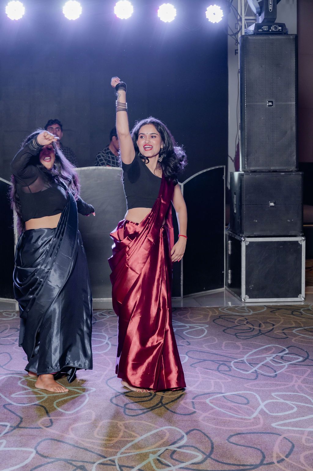 Performers dance on stage with one wearing a shimmering red sari and black crop top under dramatic stage lighting.