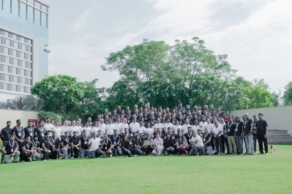 Large group of people in formal attire gathered outdoors for a panoramic photo on grassy field.
