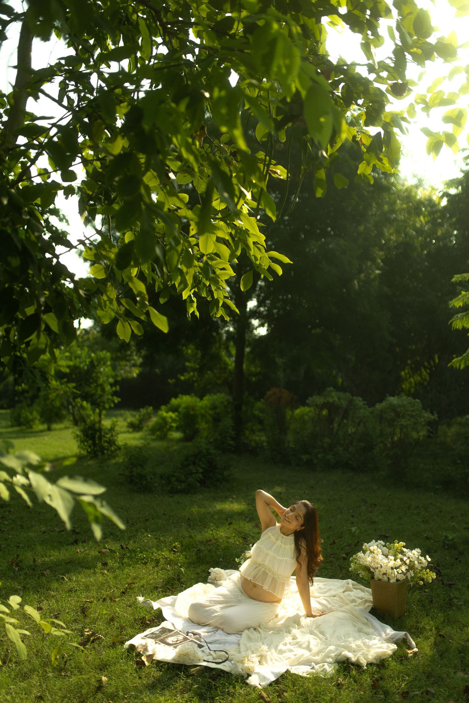 A person in a white dress reclines on a blanket in a sunlit garden surrounded by lush green foliage.