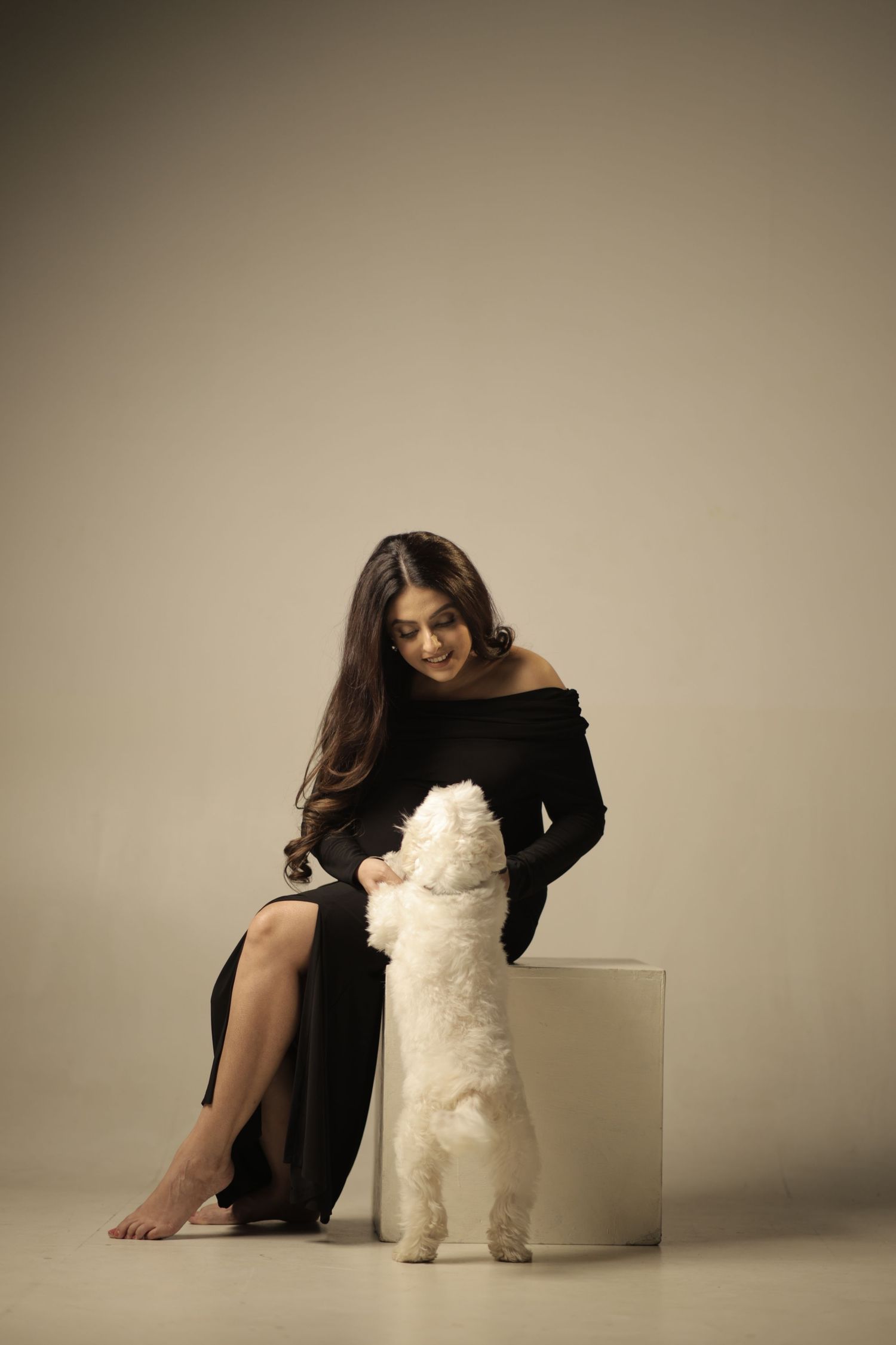 A woman in an elegant black dress sits on a white cube while interacting with a fluffy white dog standing beside her.