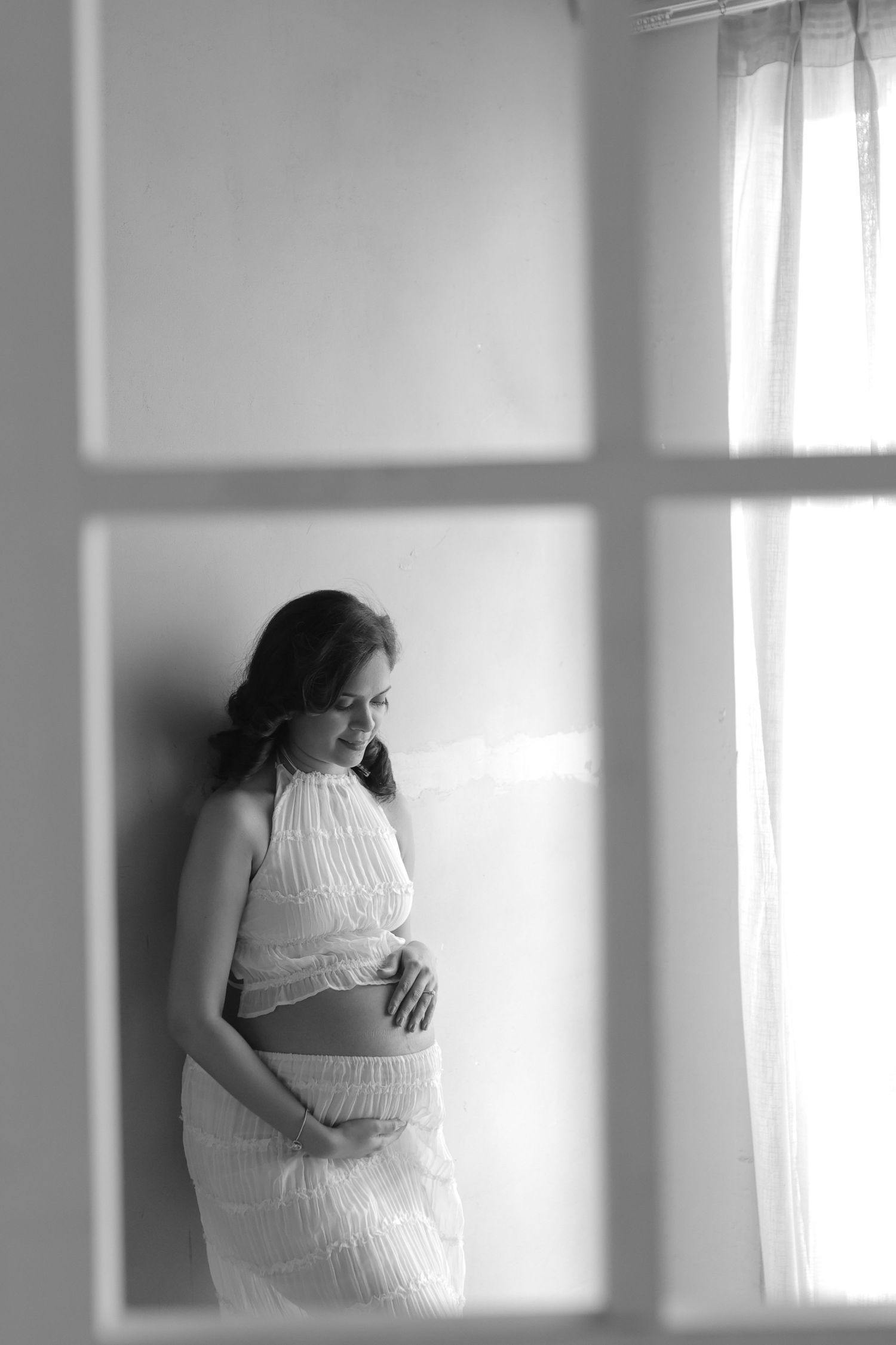 A black and white maternity photo taken through a window frame shows an expectant mother in a white dress looking down.