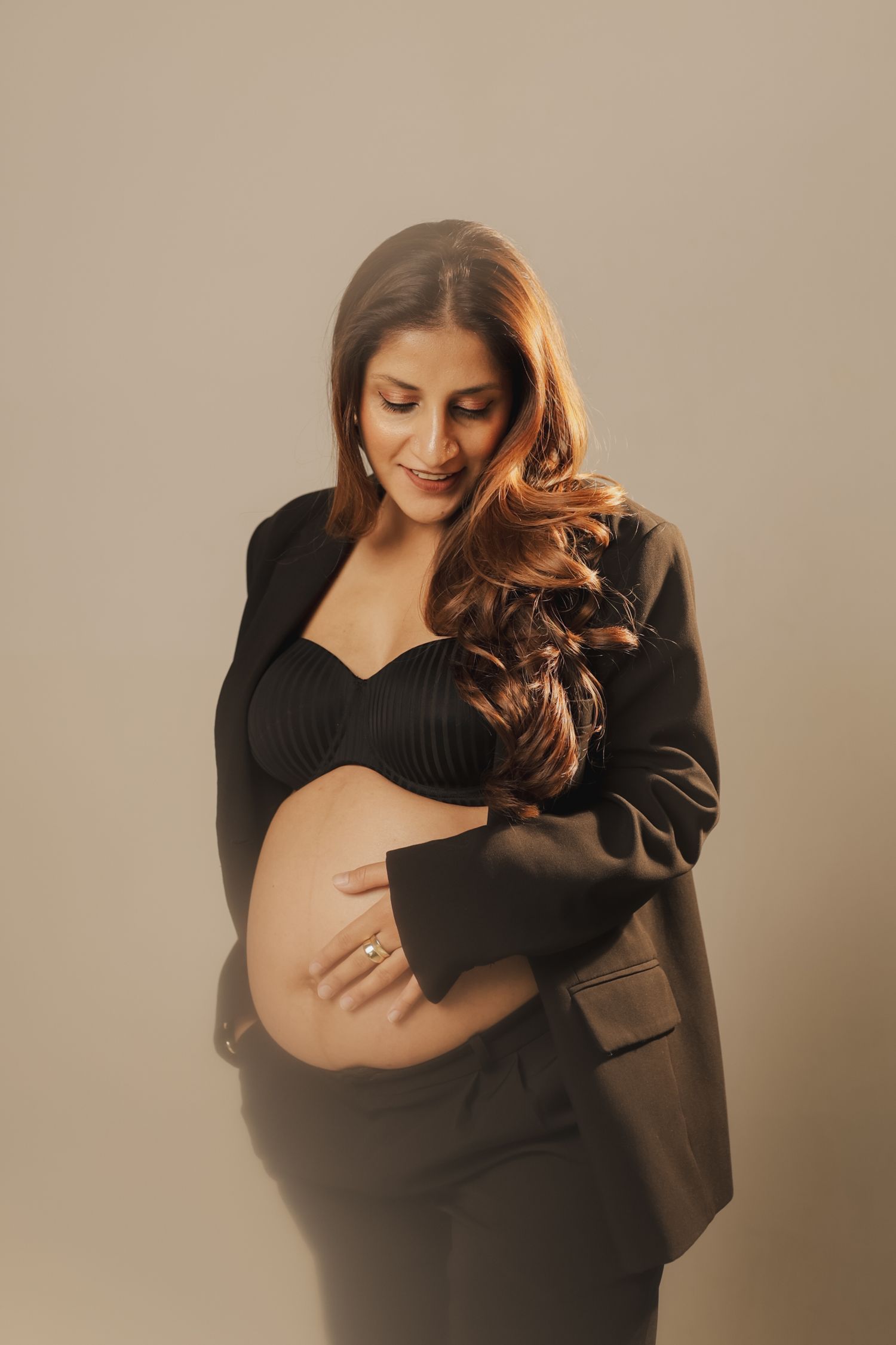 Elegant maternity portrait featuring an expectant mother in a black dress and blazer against a neutral backdrop.