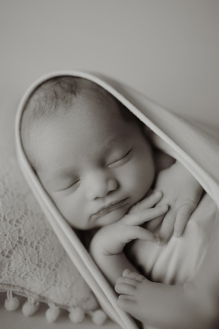 Black and white photo of a sleeping newborn baby wrapped in a soft blanket with pom-pom trim.
