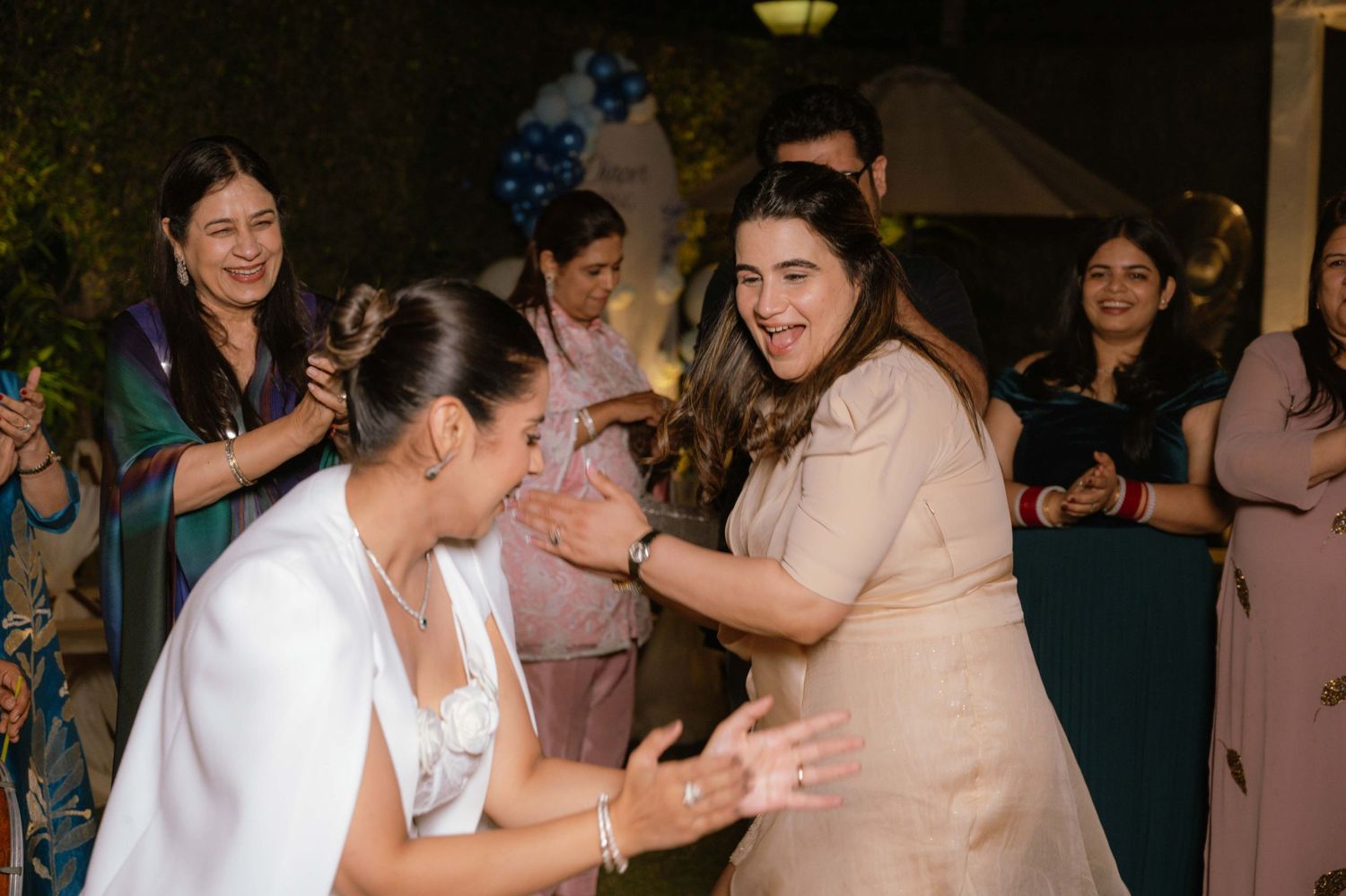 Women dancing joyfully at an outdoor wedding celebration with guests clapping in the background at night.