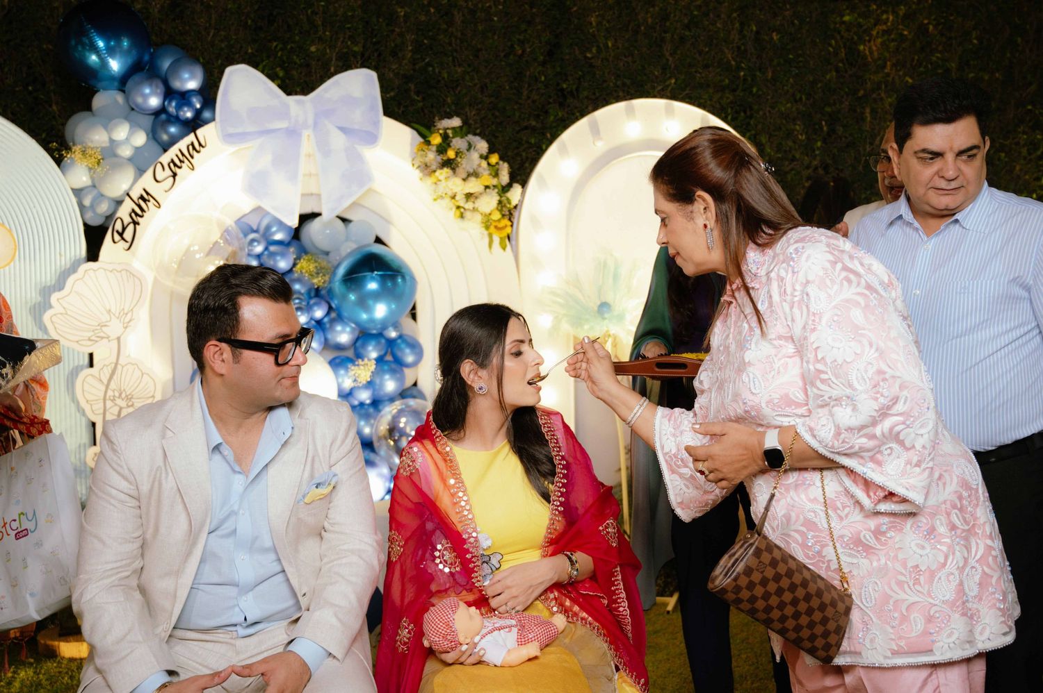A couple in festive attire sits at a baby shower decorated with blue balloons and floral backdrop while a guest in pink interacts with them.