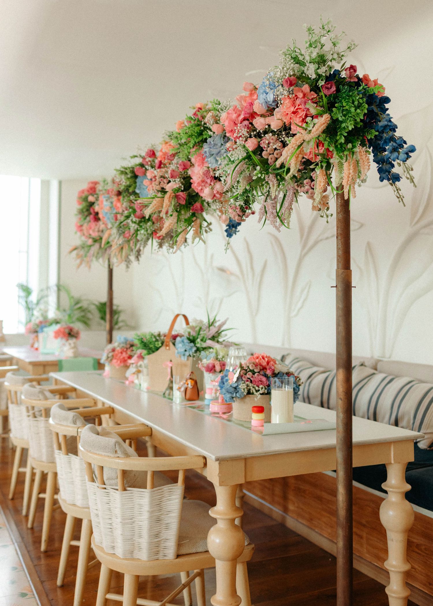 Elegant wedding reception table with wooden chairs, wicker baskets, and lush floral arch of pink, blue, and green blooms.
